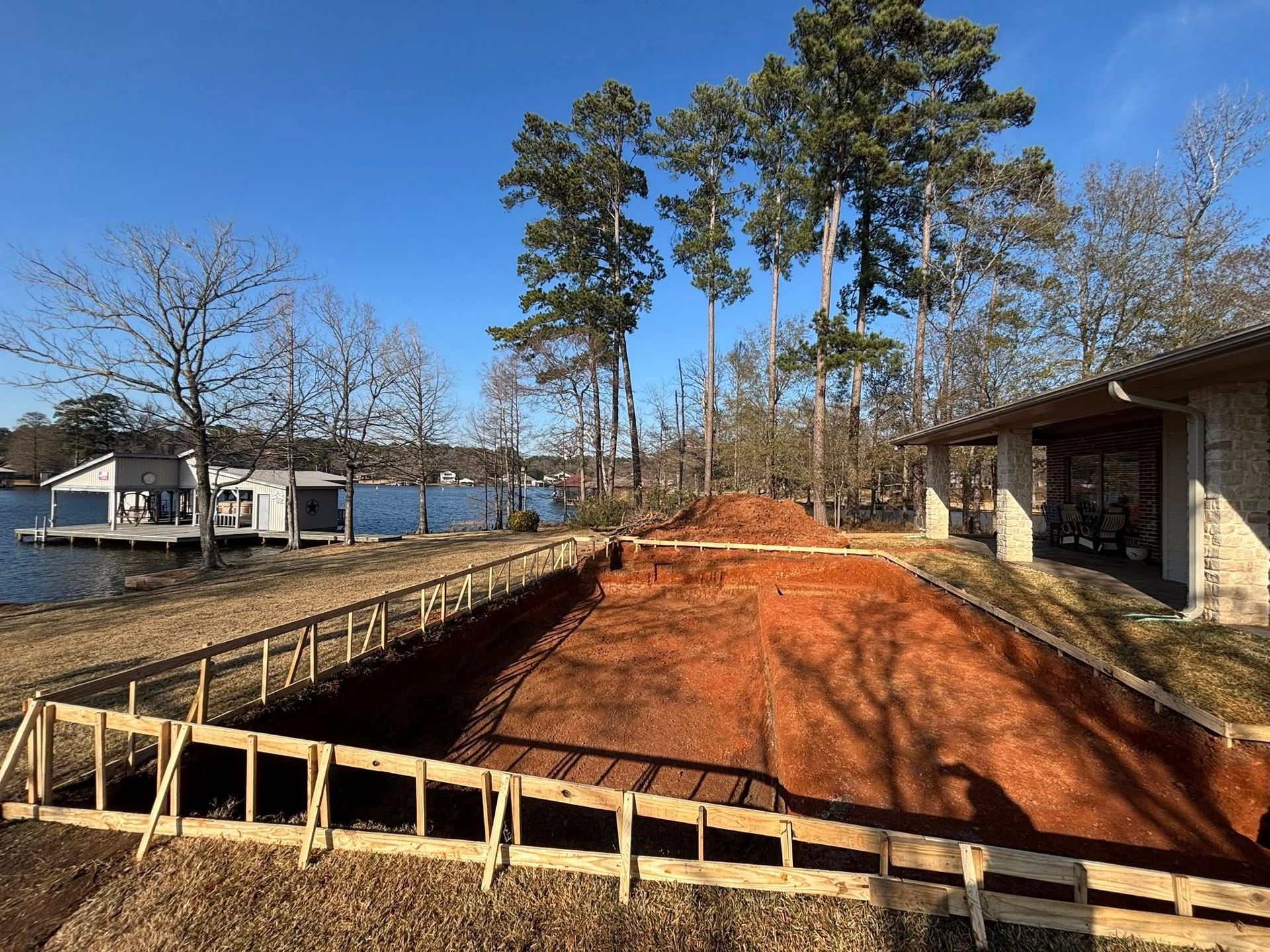 A construction site for an inground pool with red clay soil, wood framing, and a lakeside setting on a sunny day.