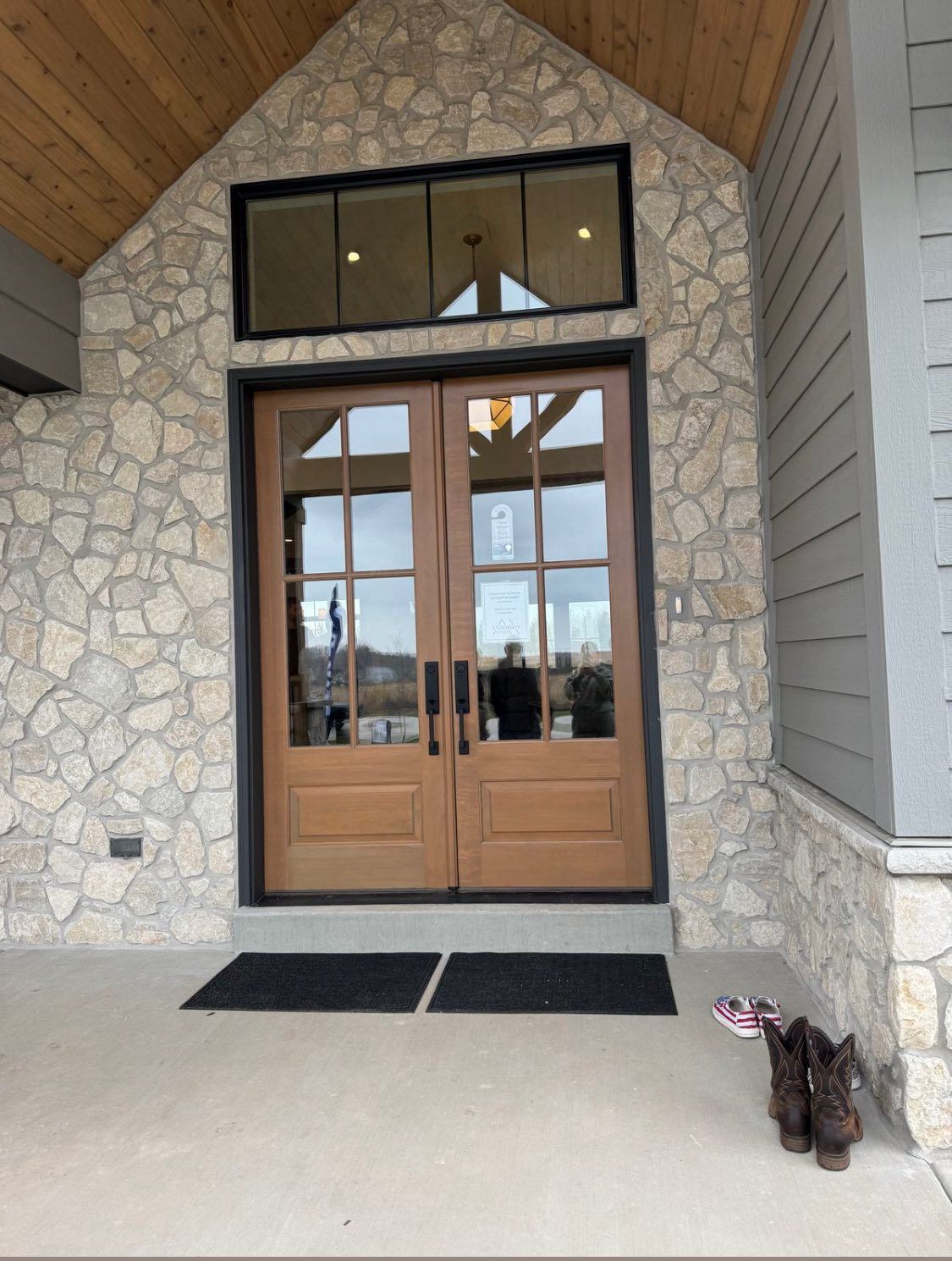 A wooden double front door with glass panels and a transom window, set in a light stone wall with a concrete porch.
