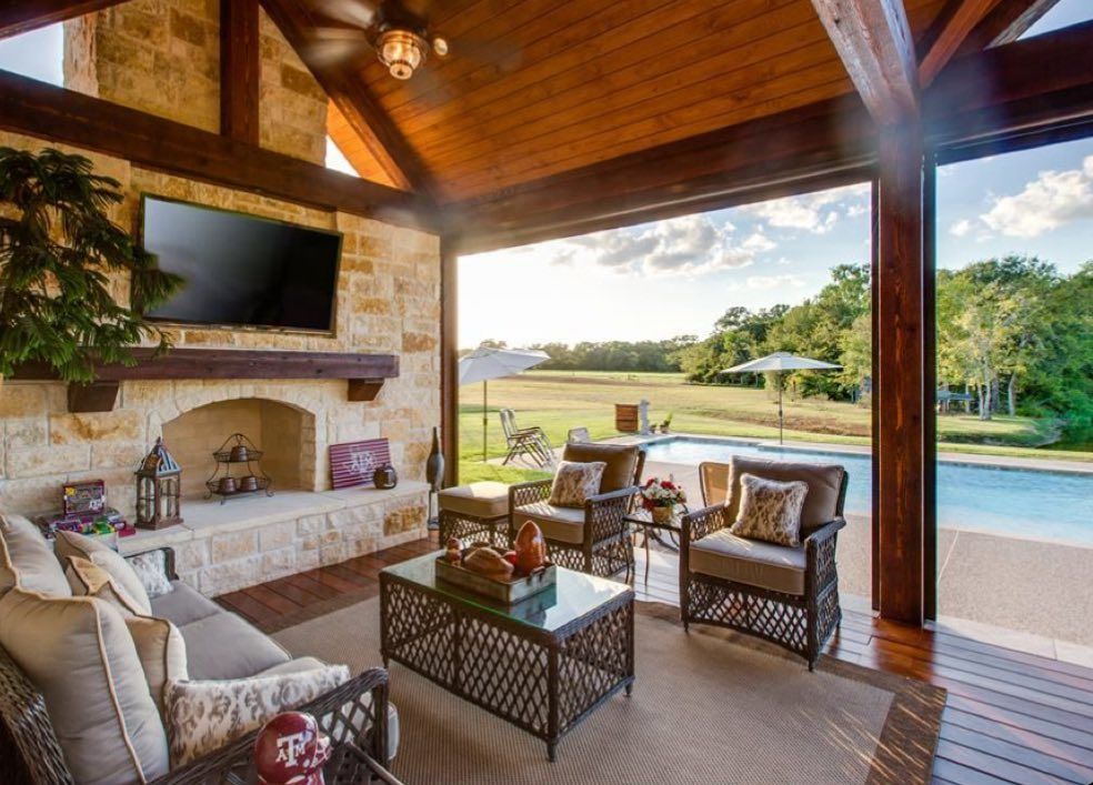 A covered outdoor living room with a stone fireplace, TV, and seating overlooking a swimming pool and grassy field.