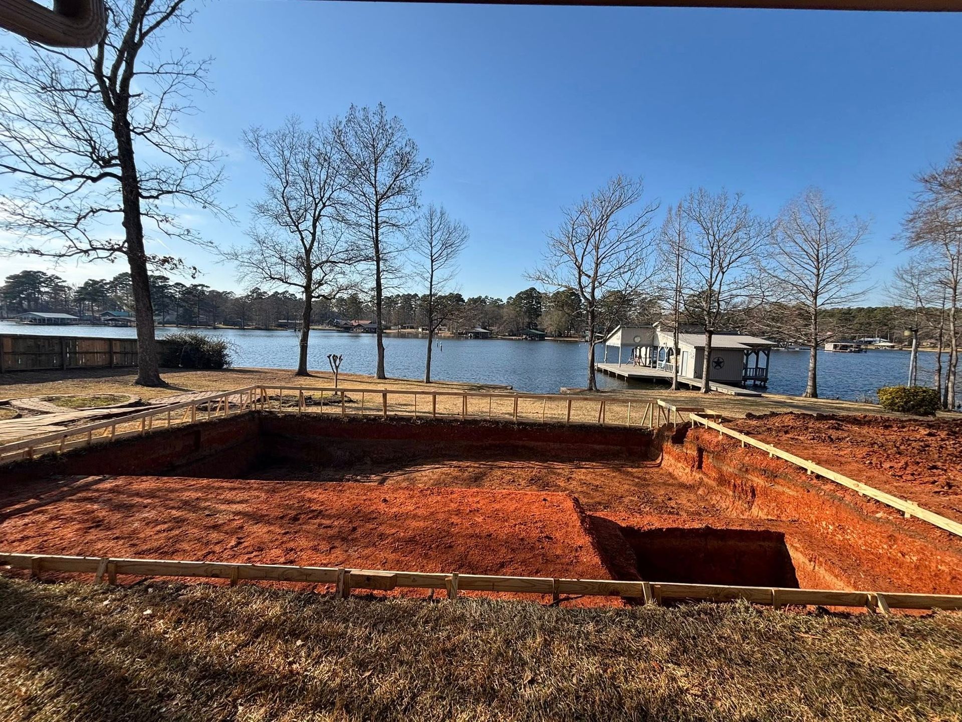 A rectangular pool foundation dug into red clay soil on a lakeshore, framed by wooden boards under a clear blue sky.