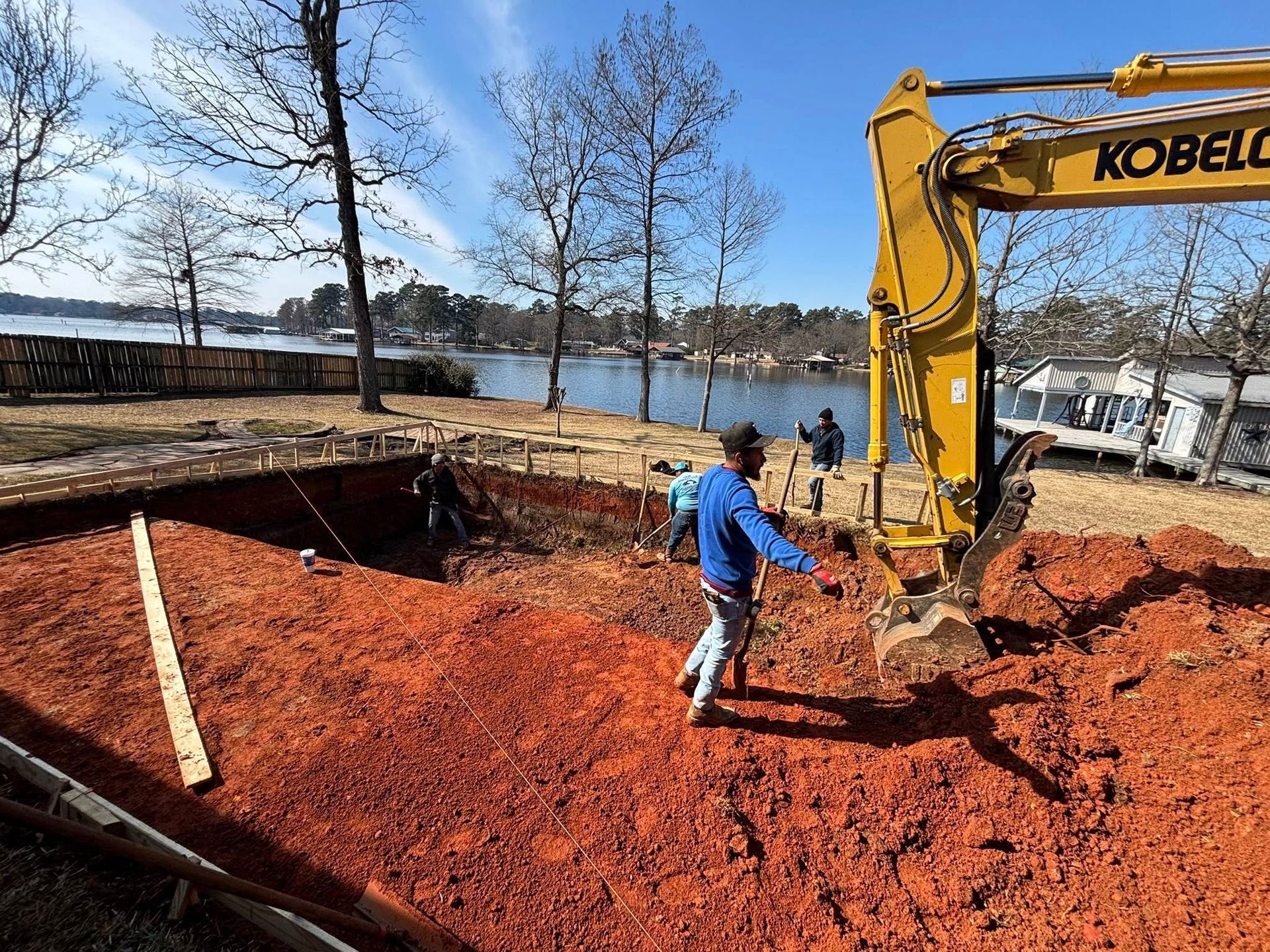 A yellow Kobelco excavator working at a lakeside construction site with red soil and workers nearby.