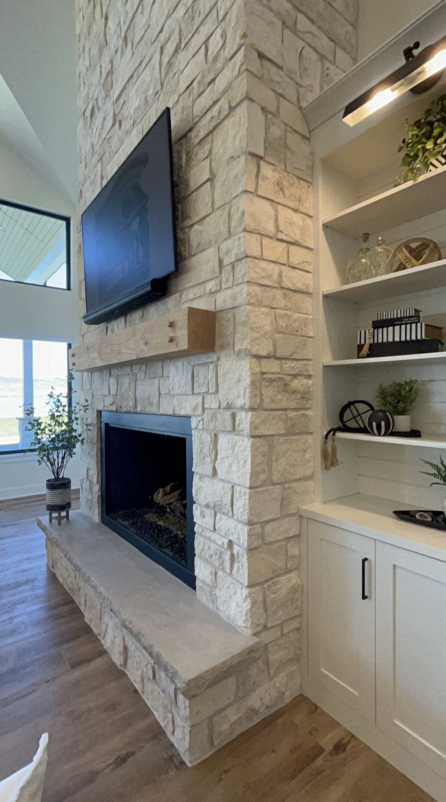 A floor-to-ceiling cream-colored stone fireplace with a wood mantel, mounted TV, and white built-in shelving beside it.