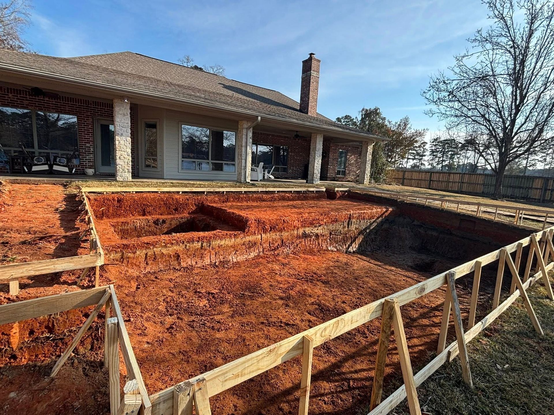 A partially excavated rectangular swimming pool site with wooden forms and orange clay soil in front of a stone house.