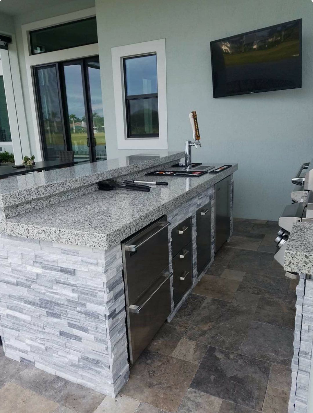An outdoor kitchen with granite countertops, stone veneer base, stainless appliances, and a mounted flat-screen TV.