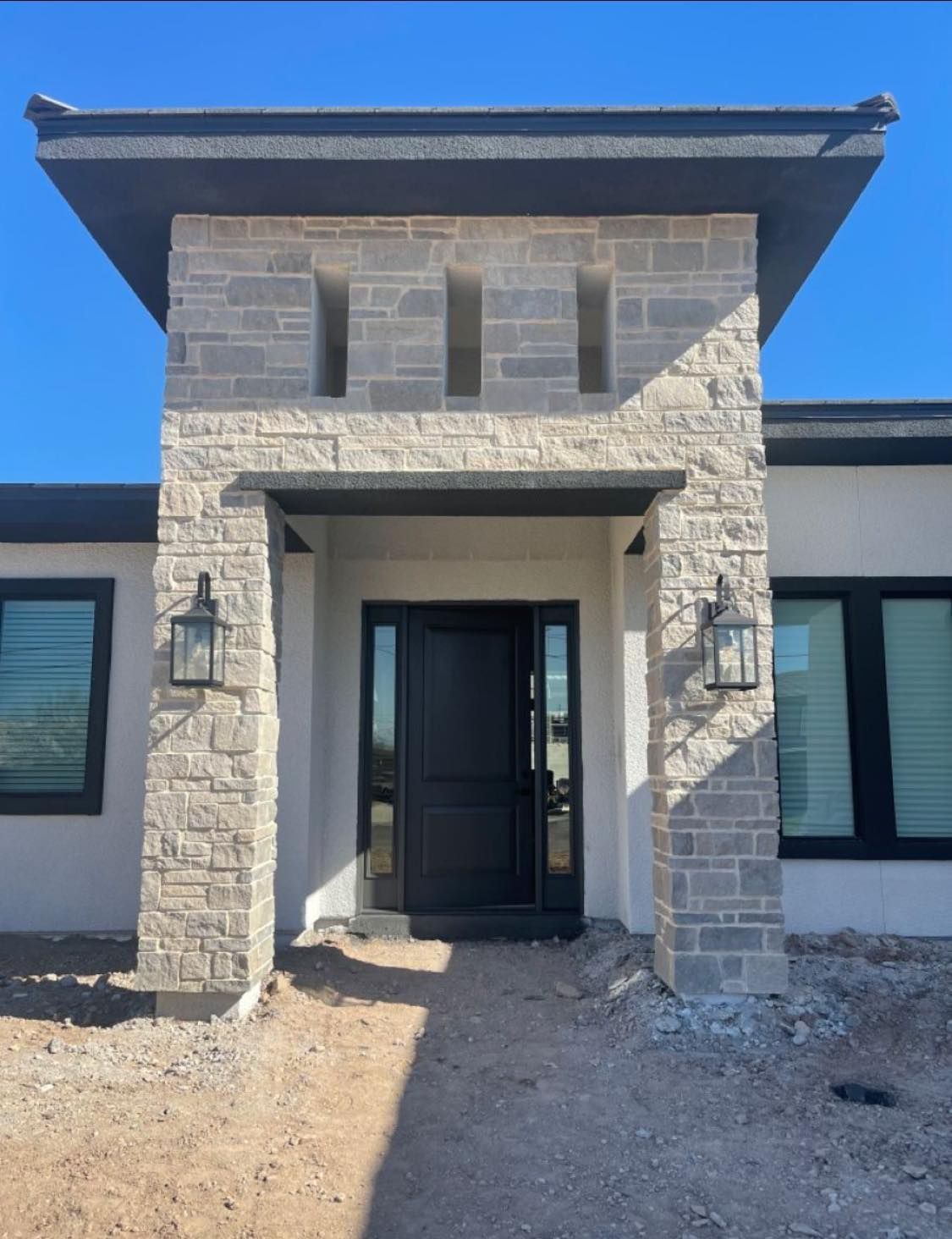 Modern house entrance with a stone-accented facade, dark door, and black window frames under a clear blue sky.