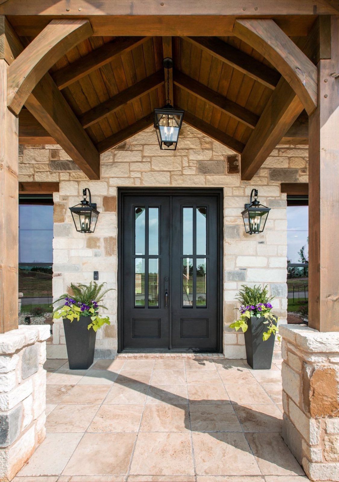 A stone-fronted entryway with a dark double door, hanging lanterns, and two potted plants under a wooden gable roof.