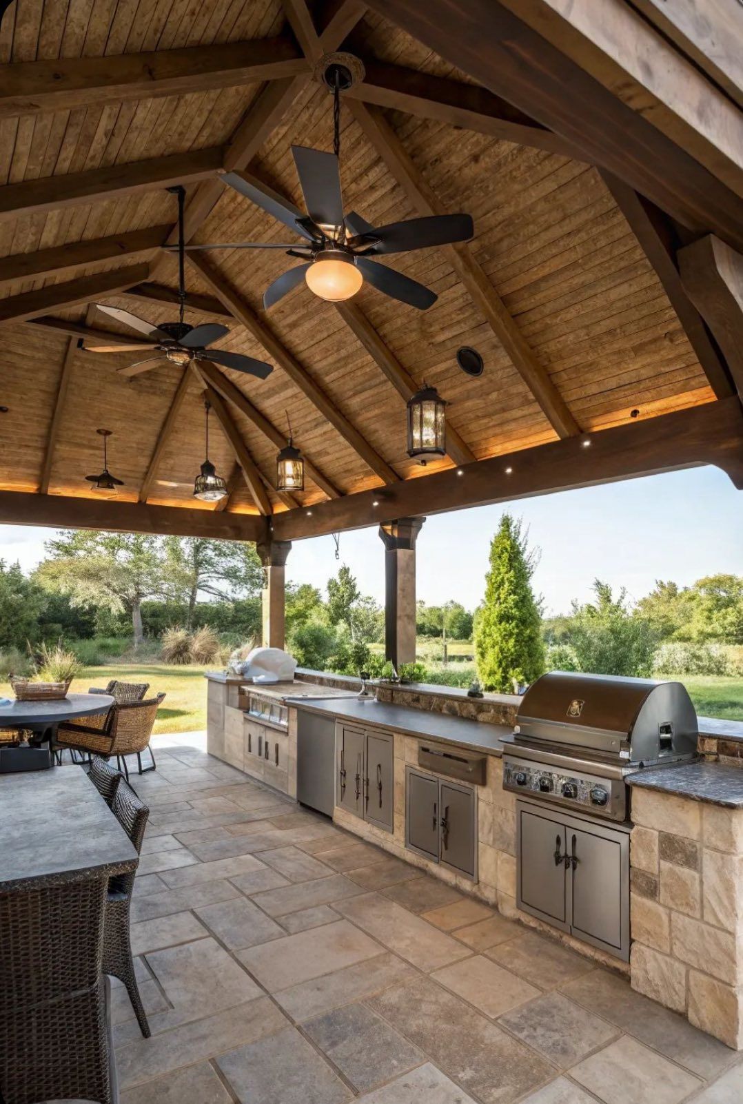 An outdoor kitchen with stone counters, stainless steel appliances, and ceiling fans under a rustic wooden pavilion.