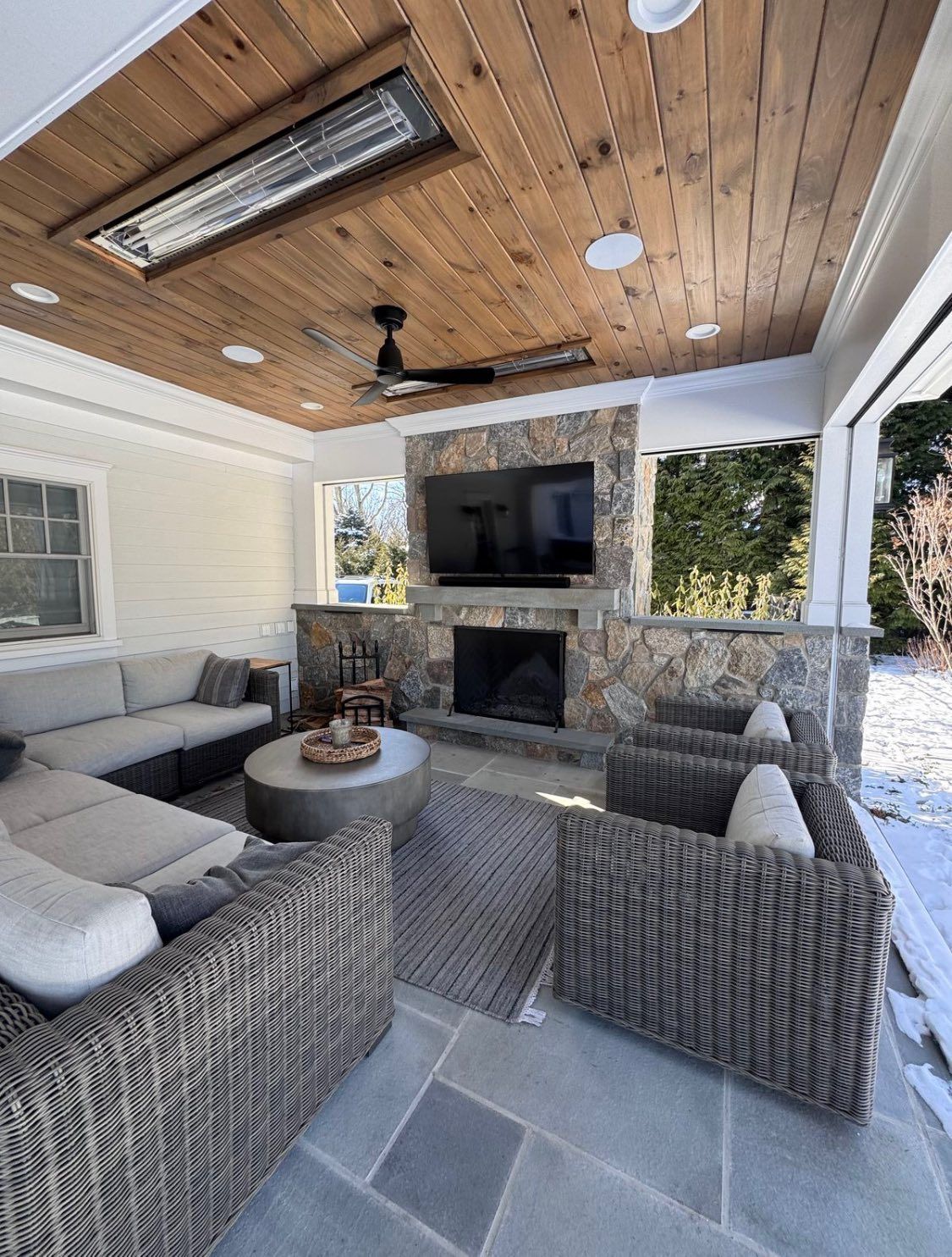 A covered outdoor patio with a stone fireplace, a wall-mounted TV, grey wicker seating, and a wood-paneled ceiling.