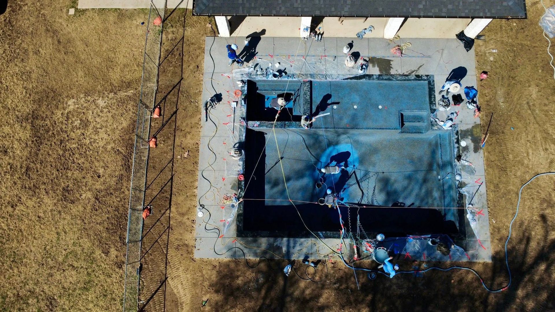 An aerial view of a construction crew installing a dark-lined backyard swimming pool on a sunny day.