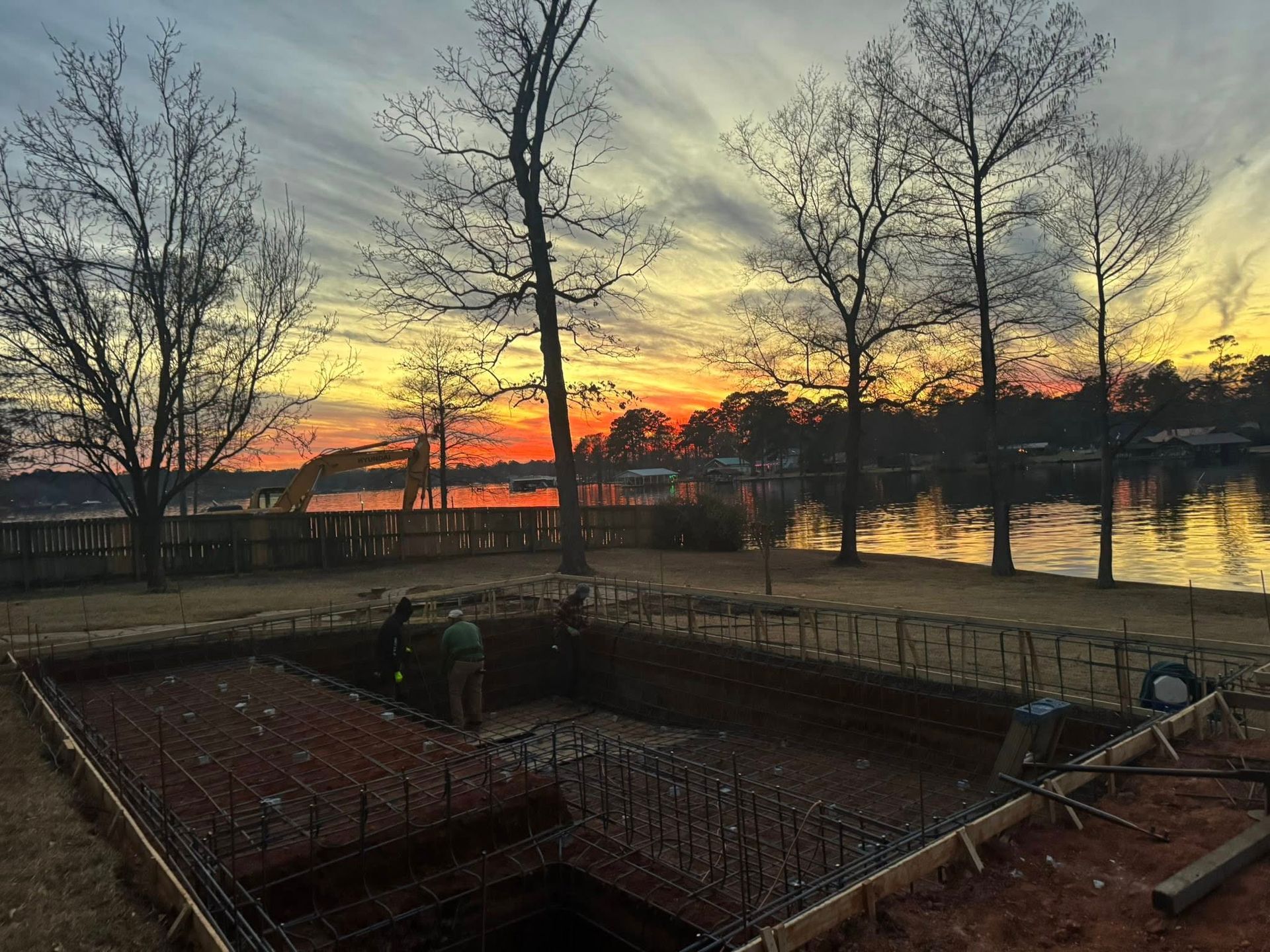 A rectangular foundation with metal rebar for a pool construction, set against a vibrant sunset by a lake.