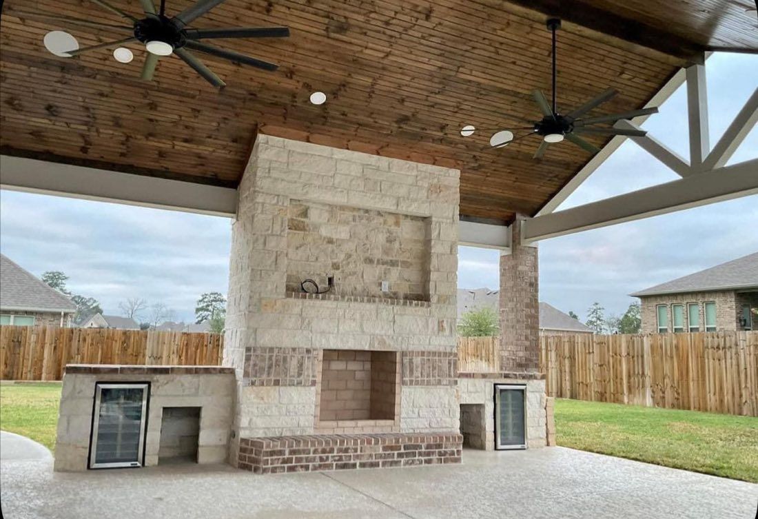 An outdoor covered patio featuring a stone fireplace, built-in counter space with appliances, and wooden ceiling beams.