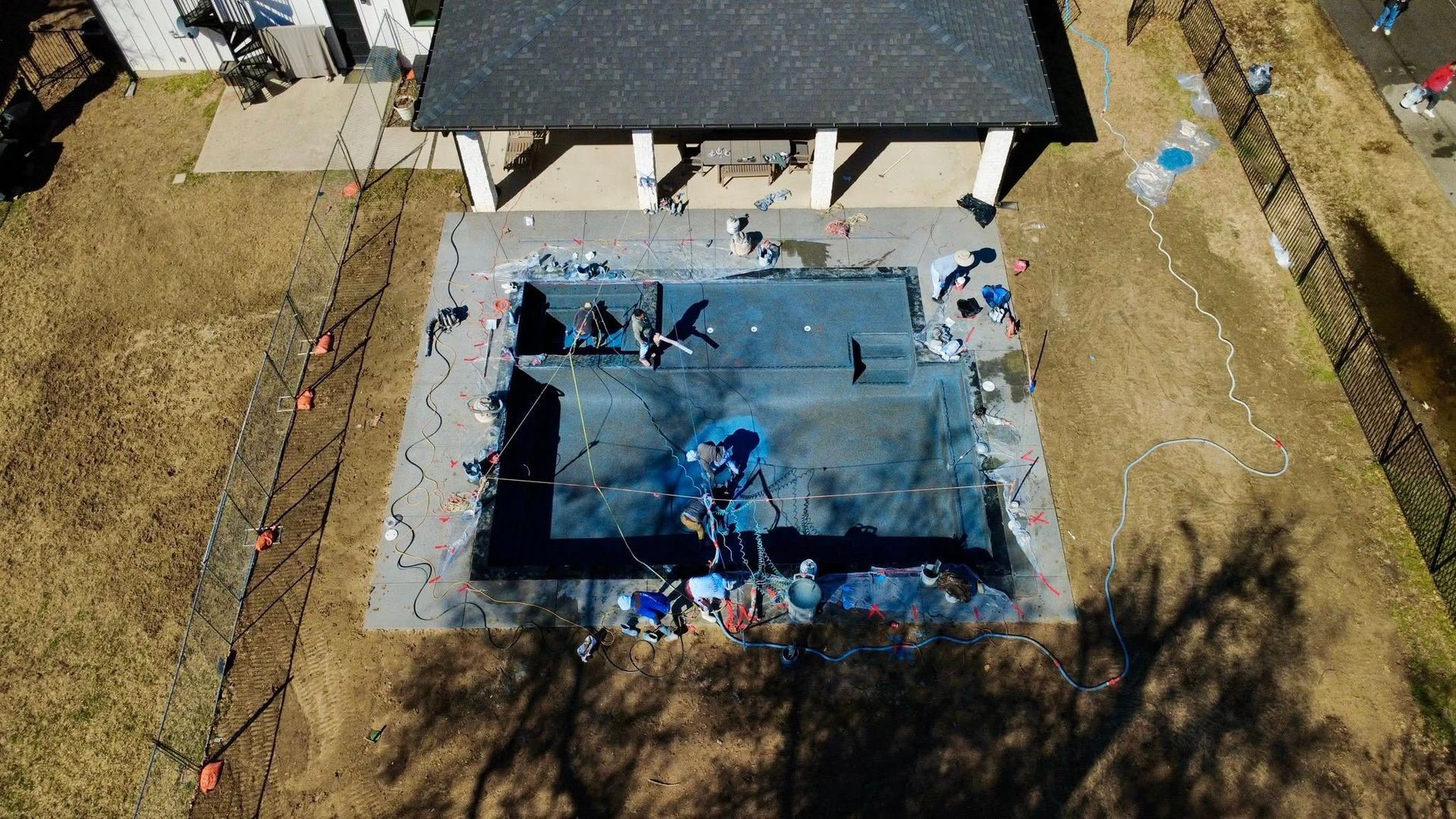 An overhead view shows construction workers repairing a rectangular swimming pool surrounded by a concrete deck.