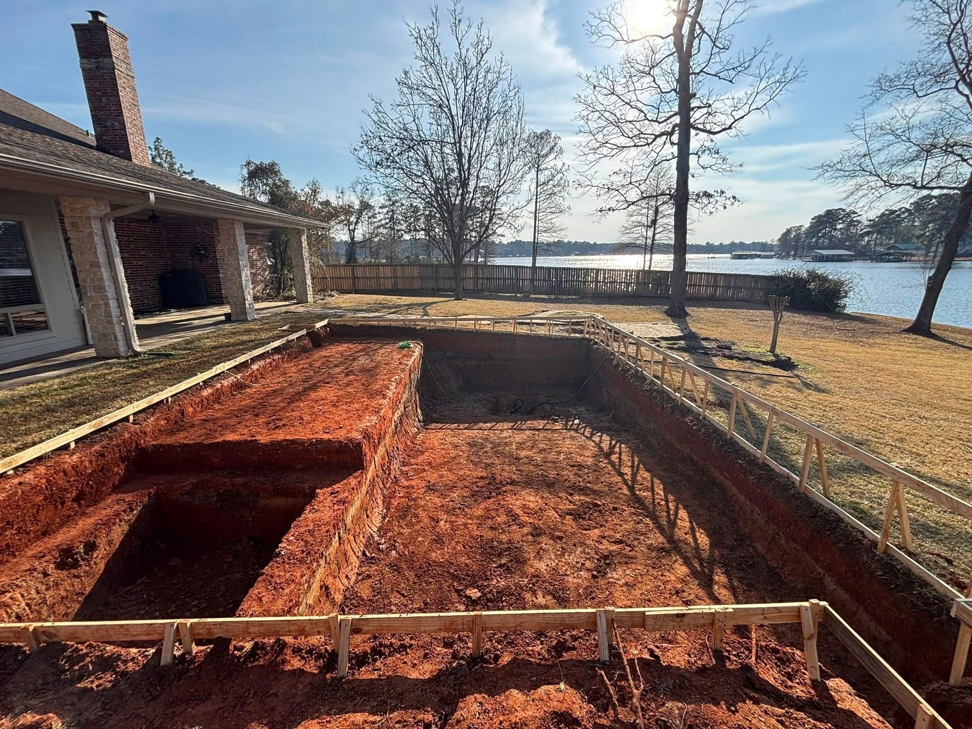 A rectangular excavation for a swimming pool in a backyard, framed by wooden boards, with a lake view in the background.