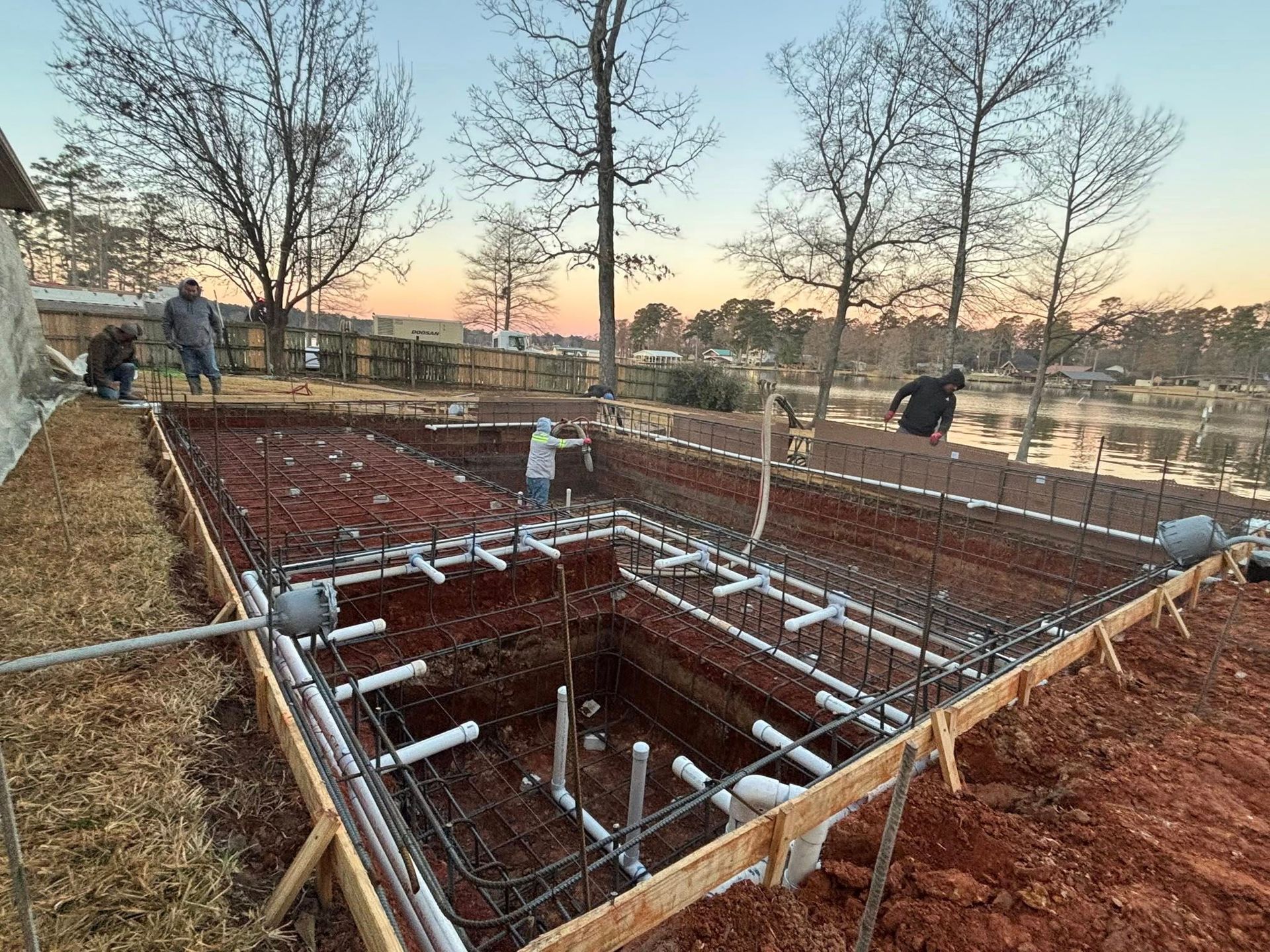 Workers assemble steel rebar and plumbing pipes in a large, excavated swimming pool foundation at a lakeside construction.