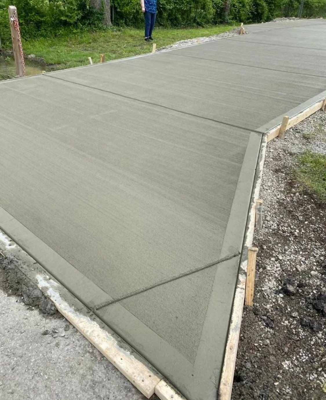A freshly poured concrete driveway section with wooden forms in place, surrounded by gravel and green grass.