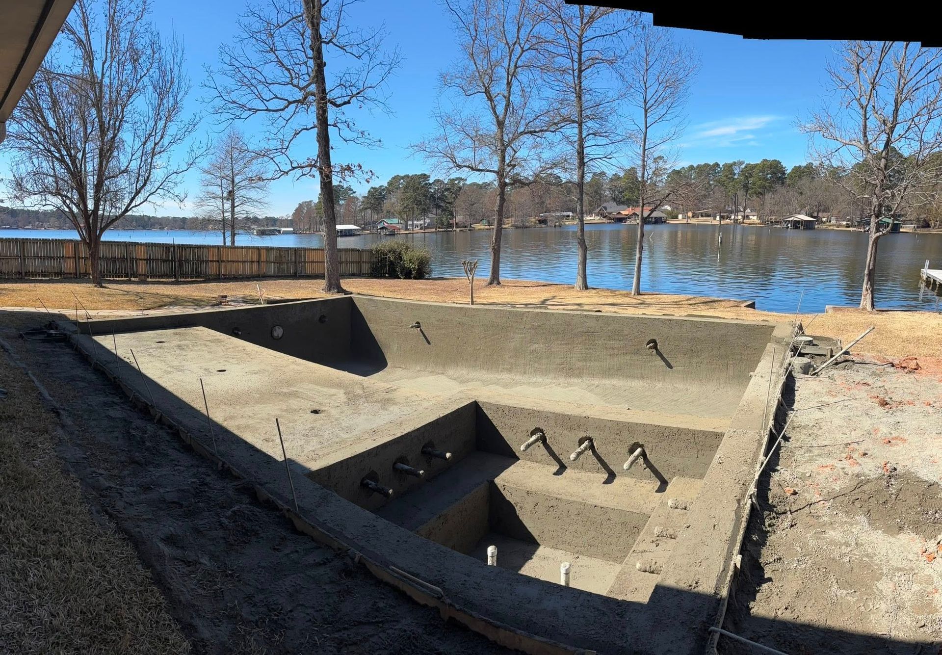 A concrete swimming pool shell under construction, featuring a tiered design overlooking a lake on a sunny day.