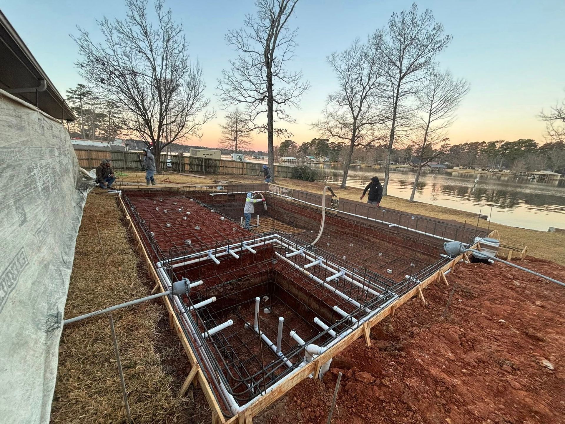 Construction crew prepares a pool foundation with rebar, piping, and wooden forms near a lake at sunset.