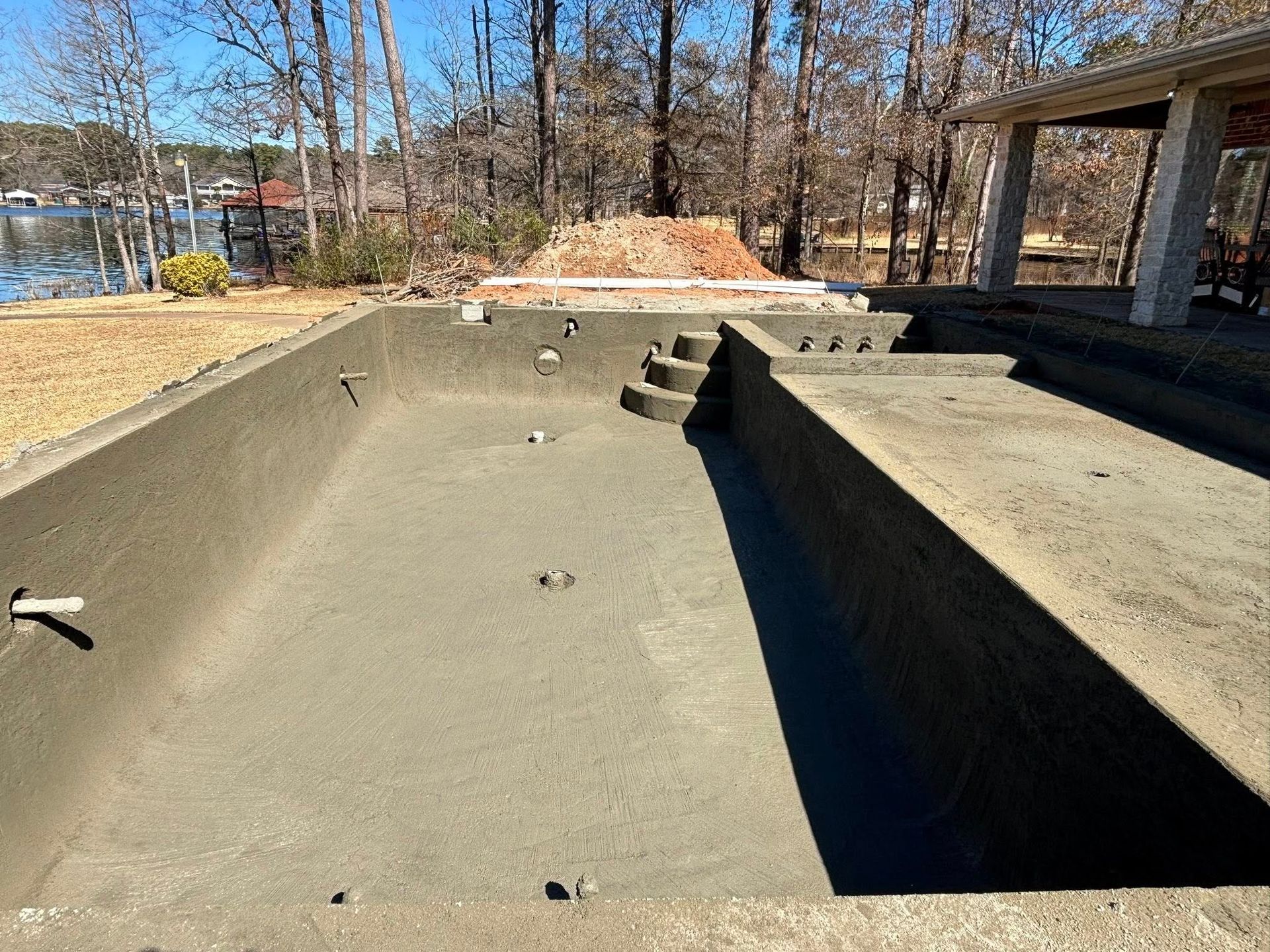 An empty, unfinished rectangular swimming pool made of concrete, situated outdoors near a house and a lake.