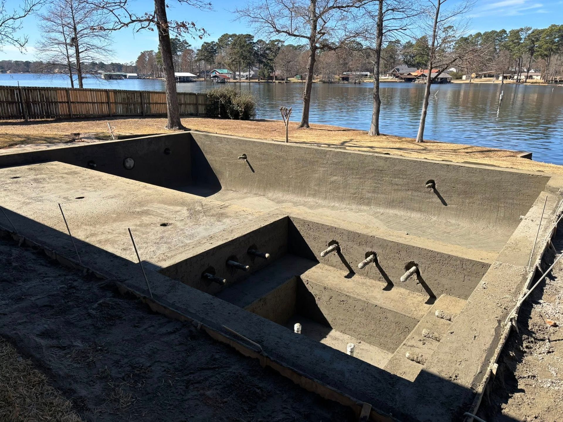 An empty, unfinished concrete swimming pool structure overlooking a lake on a sunny day.