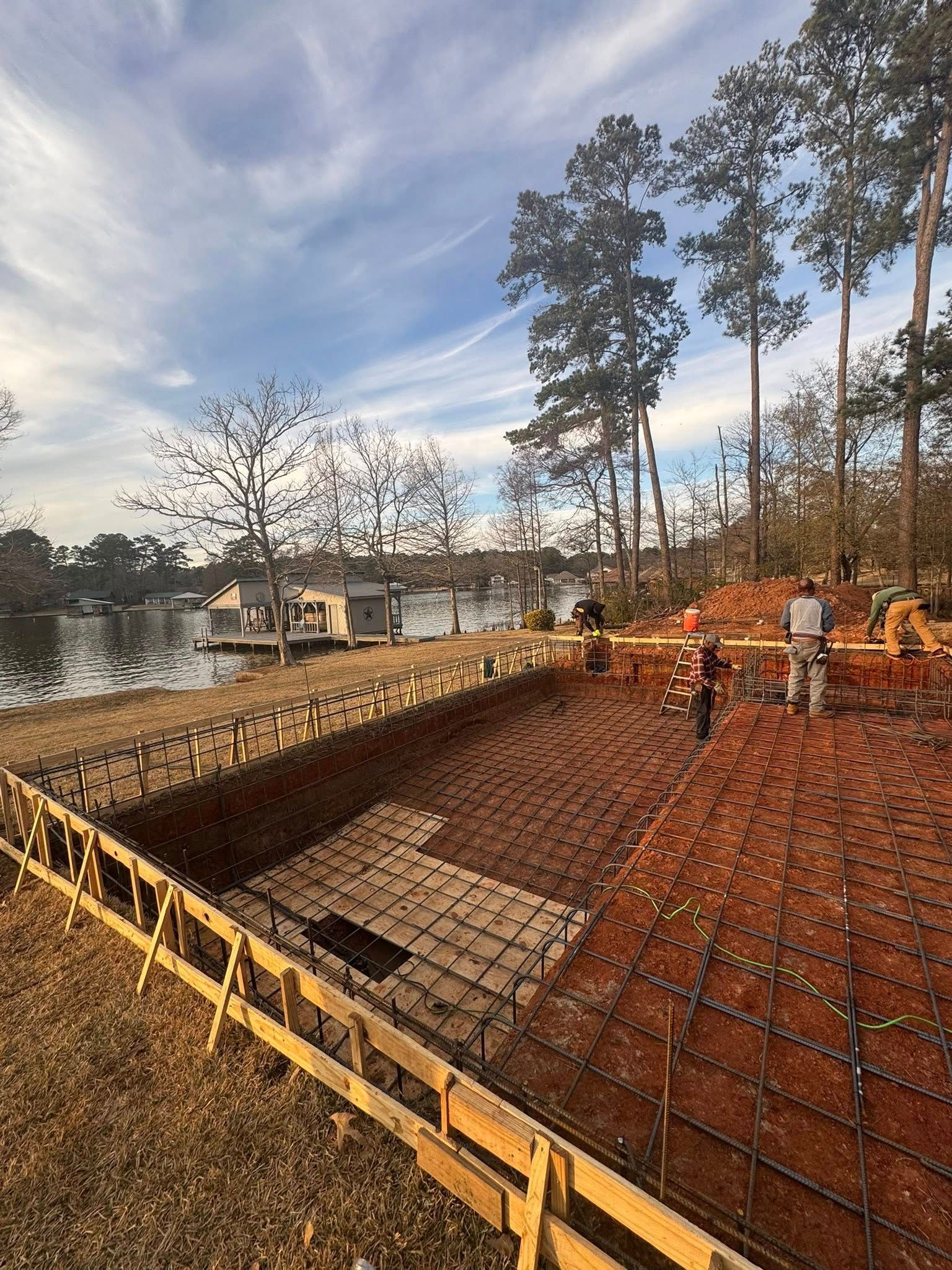 Construction workers prep a rebar-lined swimming pool excavation near a lakefront with trees under a cloudy sky.