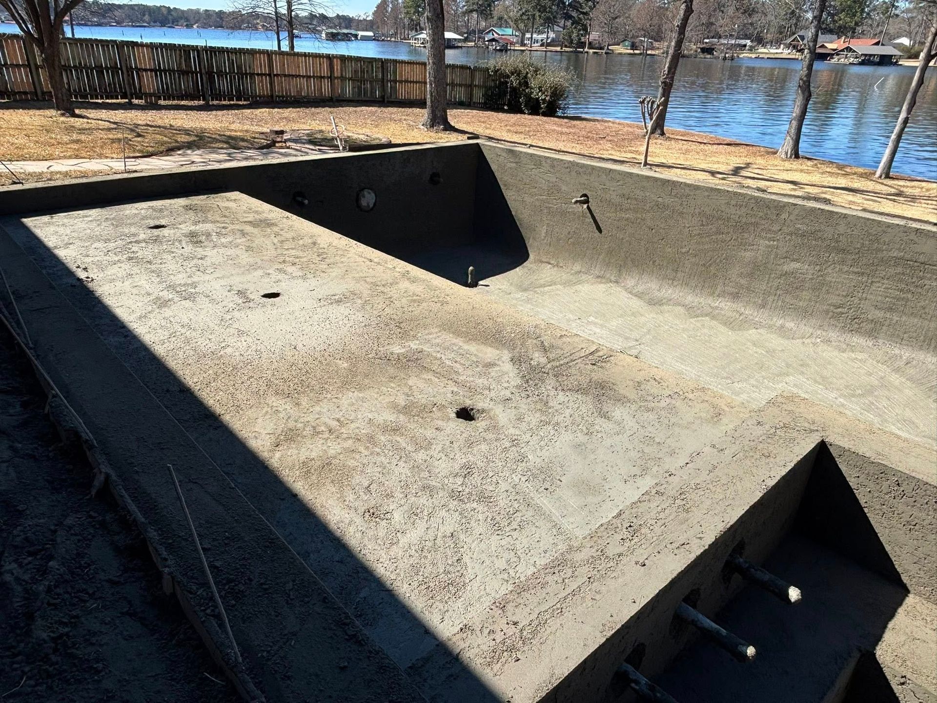 A newly constructed rectangular concrete swimming pool shell sits by a lake on a sunny day.