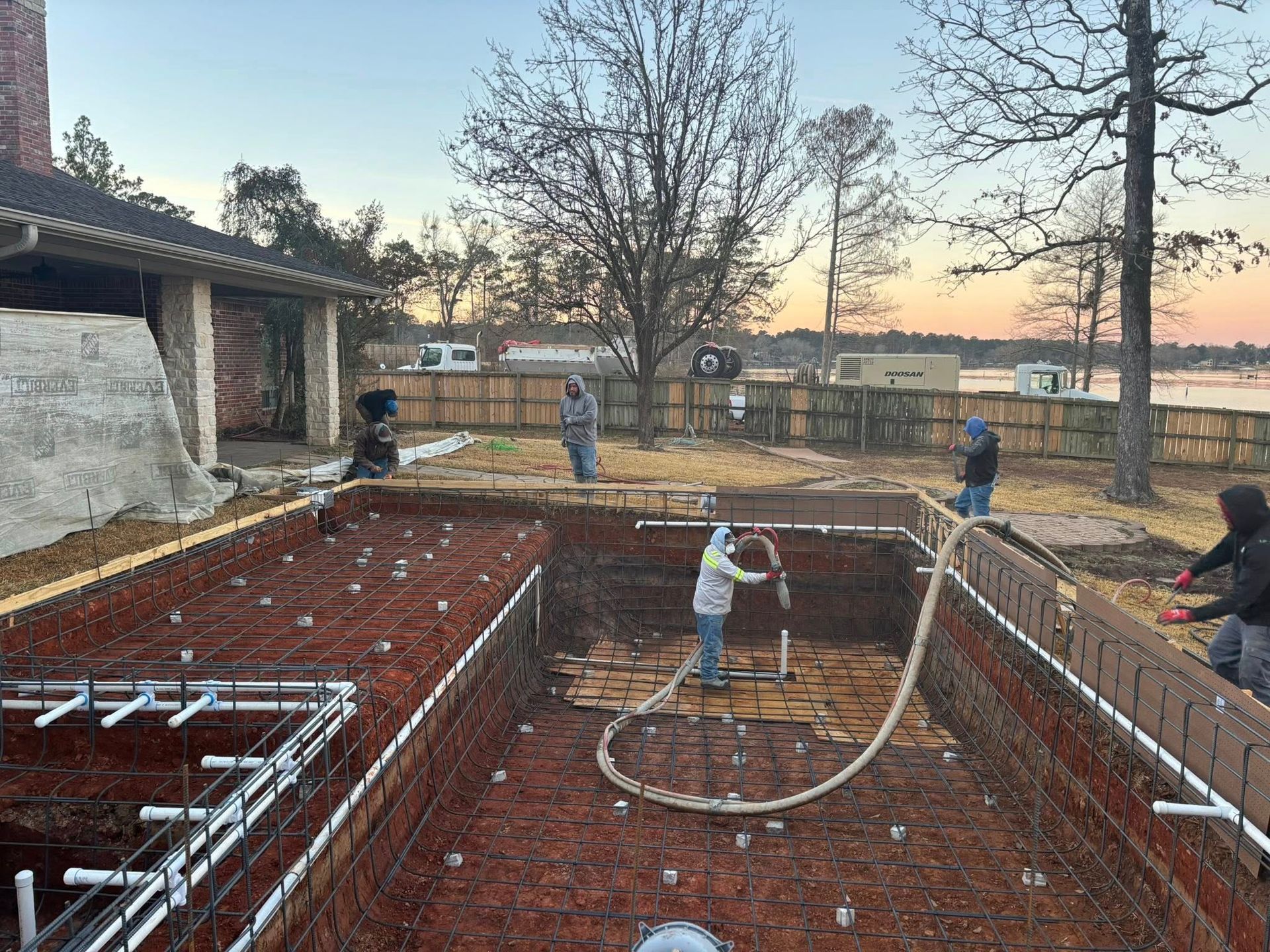 Construction crew spraying gunite into a backyard pool foundation with plumbing and steel rebar visible.
