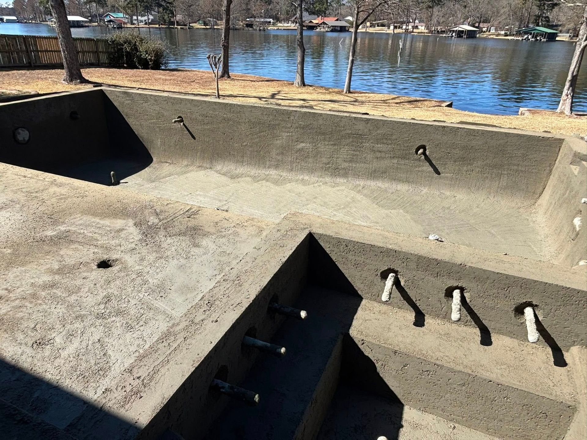 An unfinished concrete swimming pool under construction near a lake with plumbing pipes protruding from the walls.