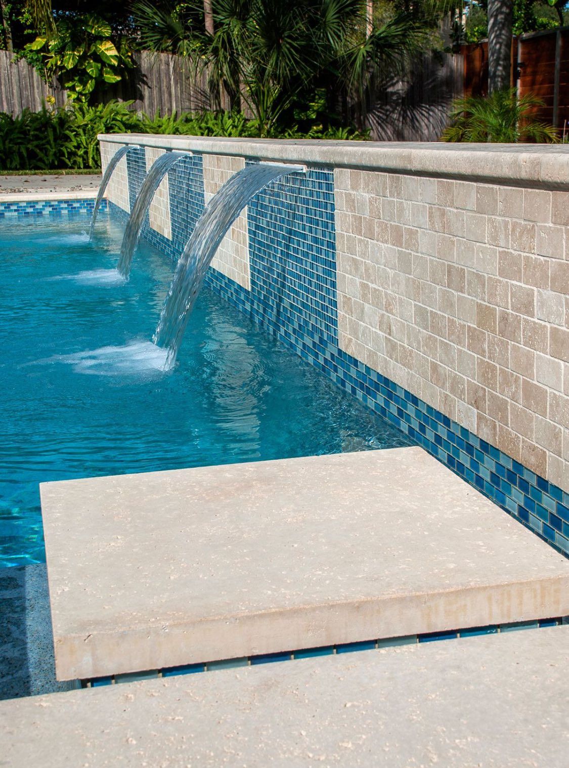 A swimming pool featuring a tan stone wall with three cascading water features and vibrant blue mosaic tile accents.