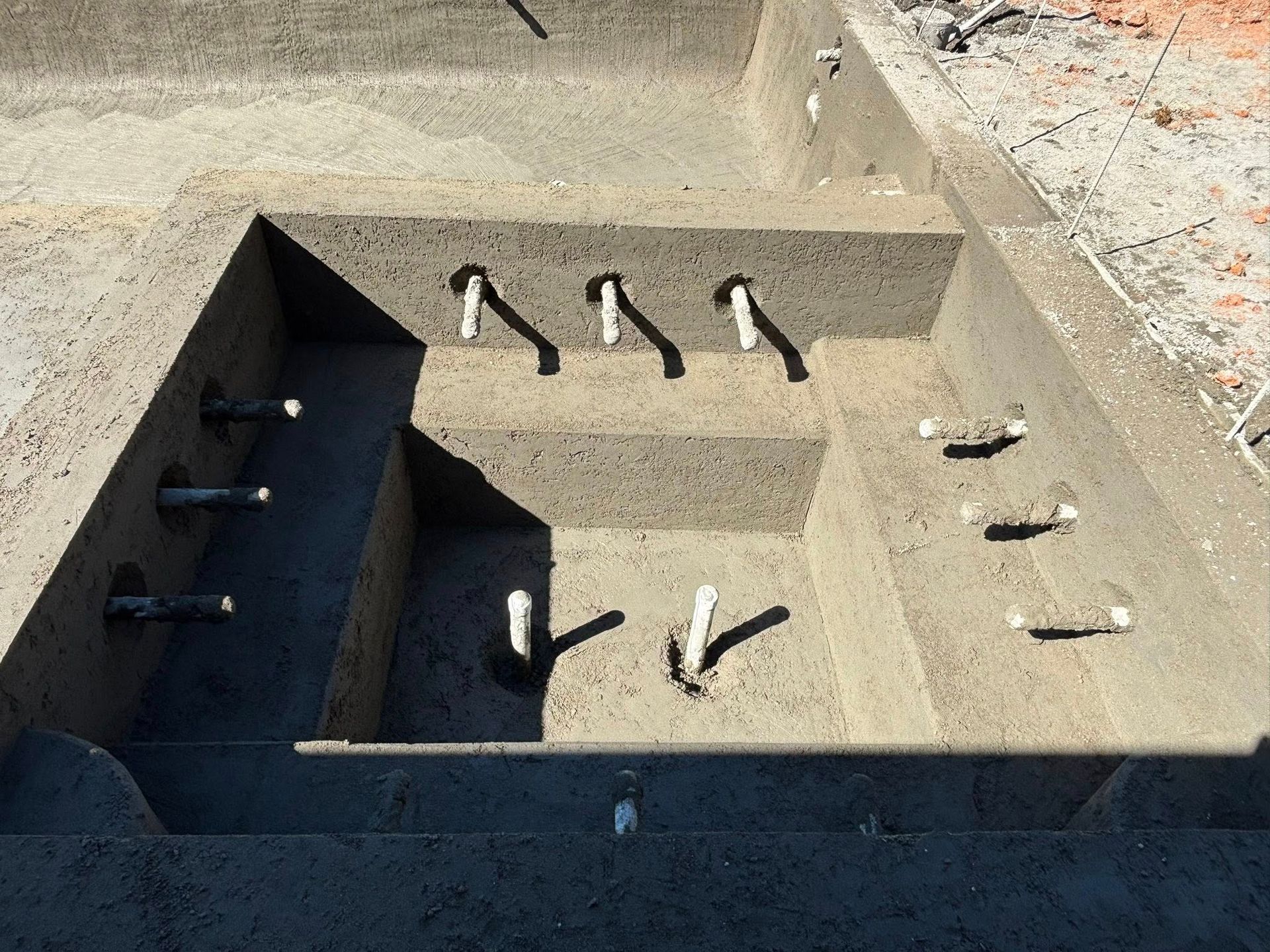 A concrete swimming pool structure under construction with several plumbing pipes protruding from the walls and steps.