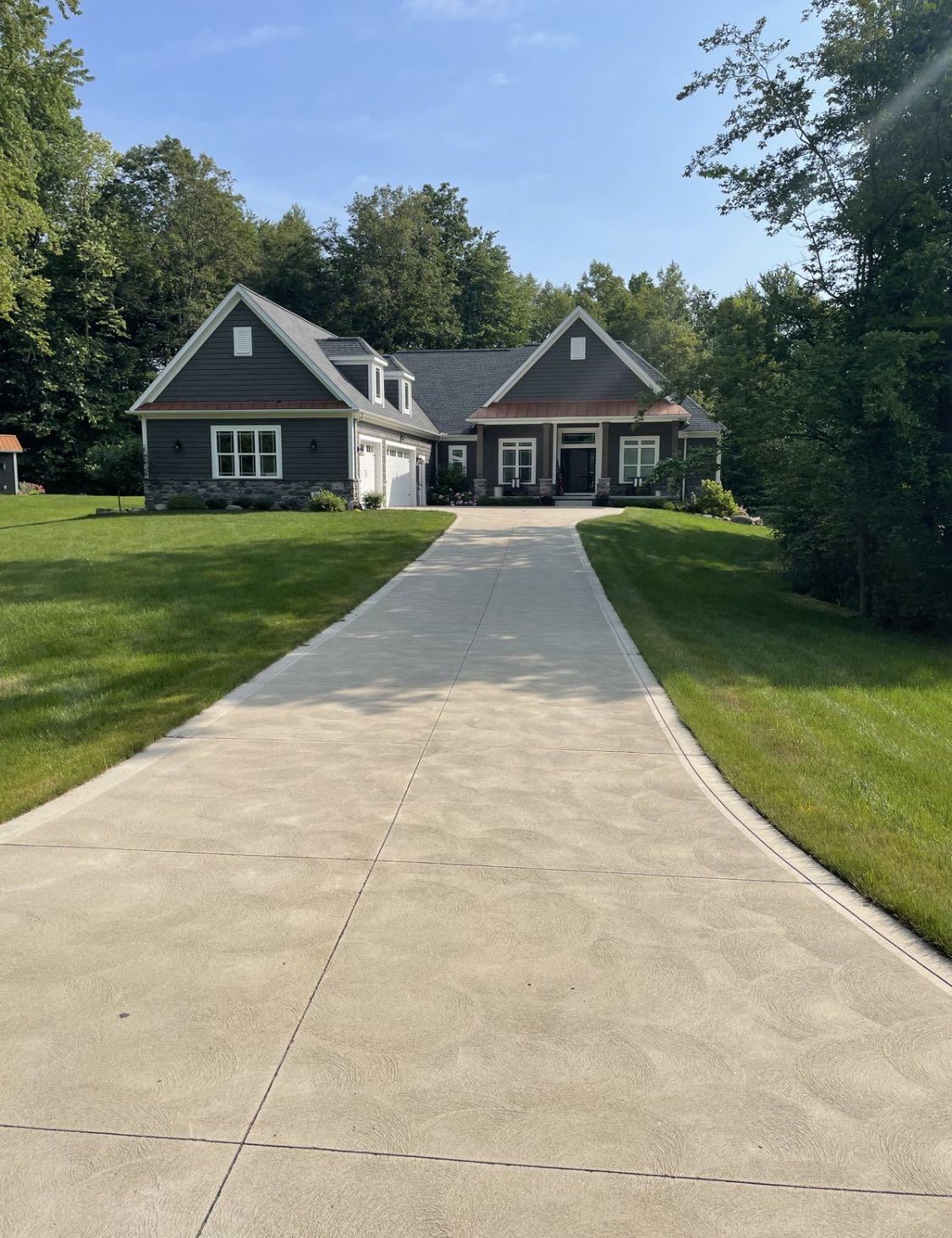 A long concrete driveway leading to a gray house with a peaked roof, surrounded by a green lawn and trees on a sunny day.