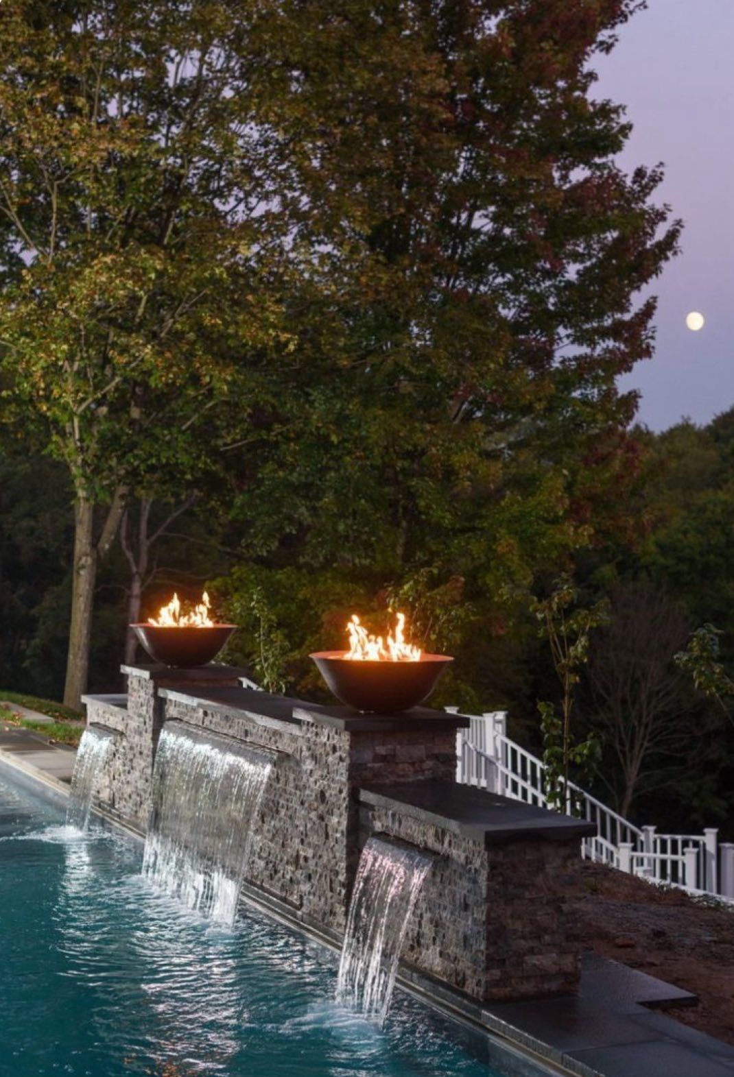 Two flaming fire bowls atop a stone water feature pouring into a swimming pool at dusk with a full moon in the background.
