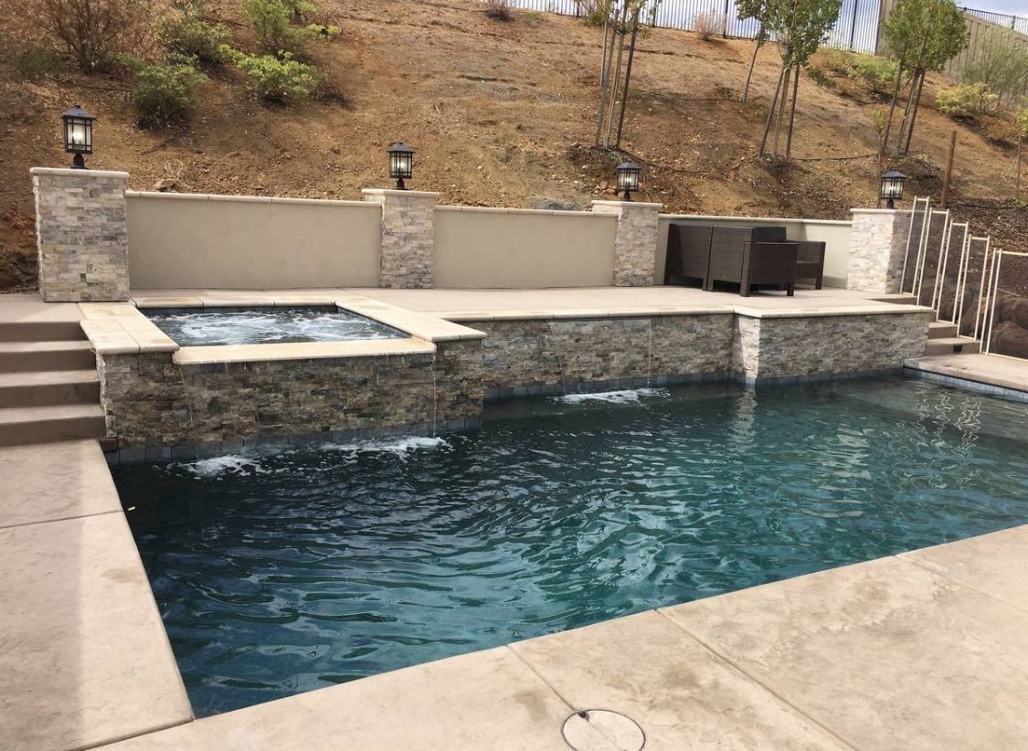 A pool with a raised stone spa and waterfall, stone pillars with lamps, and a concrete deck against a hillside backdrop.