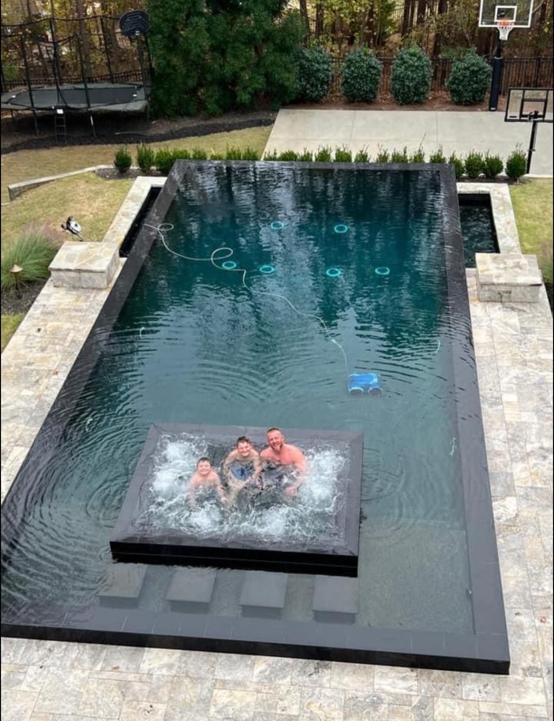 Three people sit in an elevated hot tub centered inside a dark, rectangular infinity pool in a residential backyard.