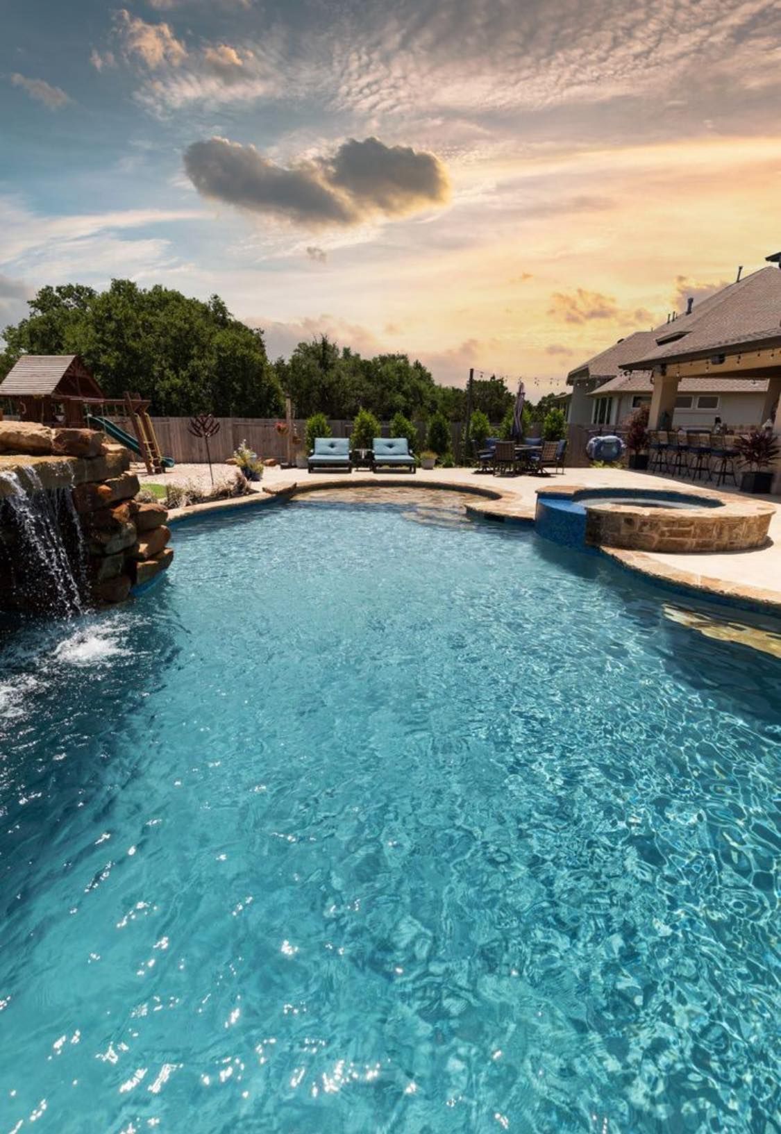 A blue backyard swimming pool with a rock waterfall feature and hot tub at sunset under a cloudy, colorful sky.