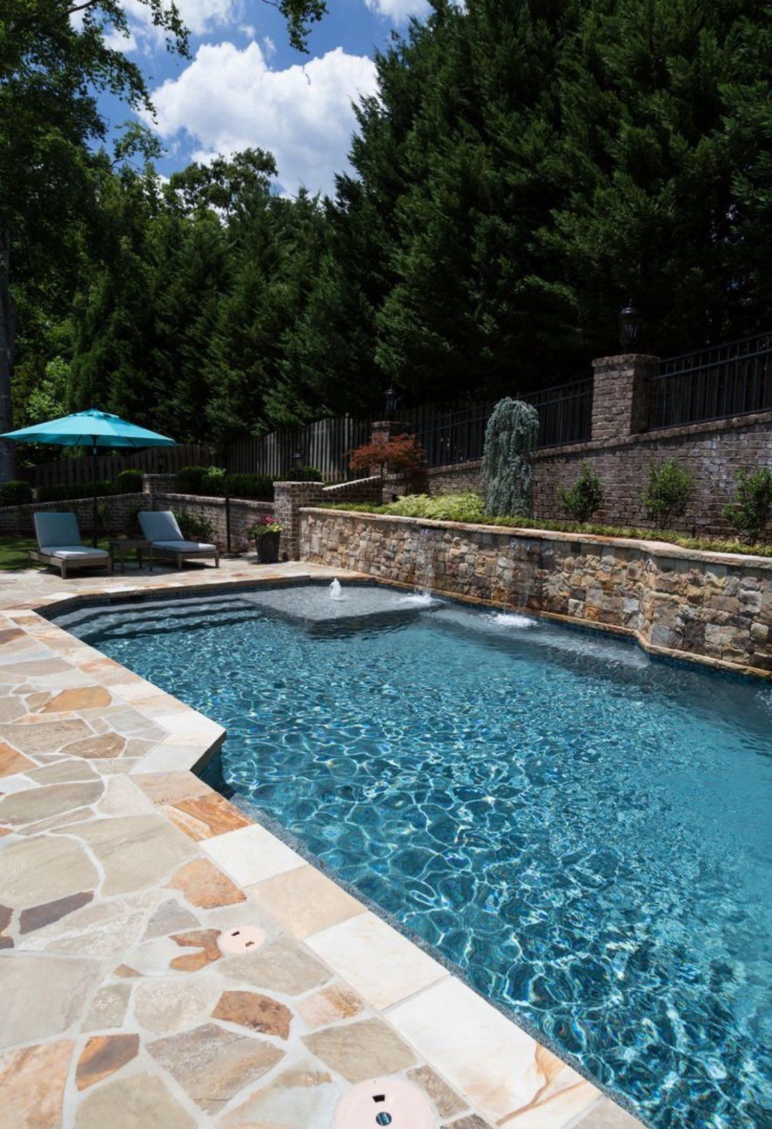 A sunlit outdoor swimming pool with a stone patio, fountains, and lounge chairs surrounded by trees and a stone wall.