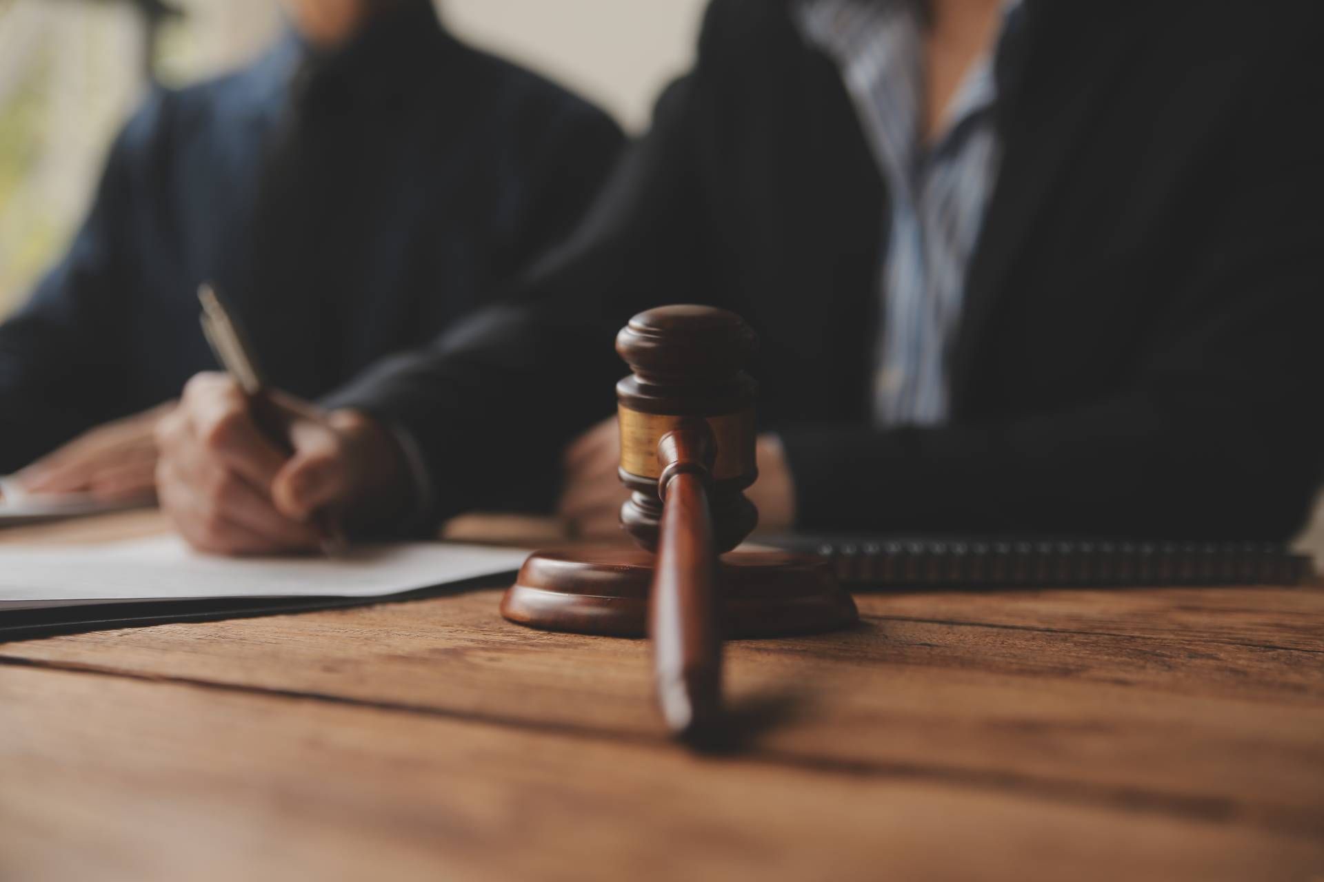 Wooden gavel on a table with two people in suits, one writing on a paper.