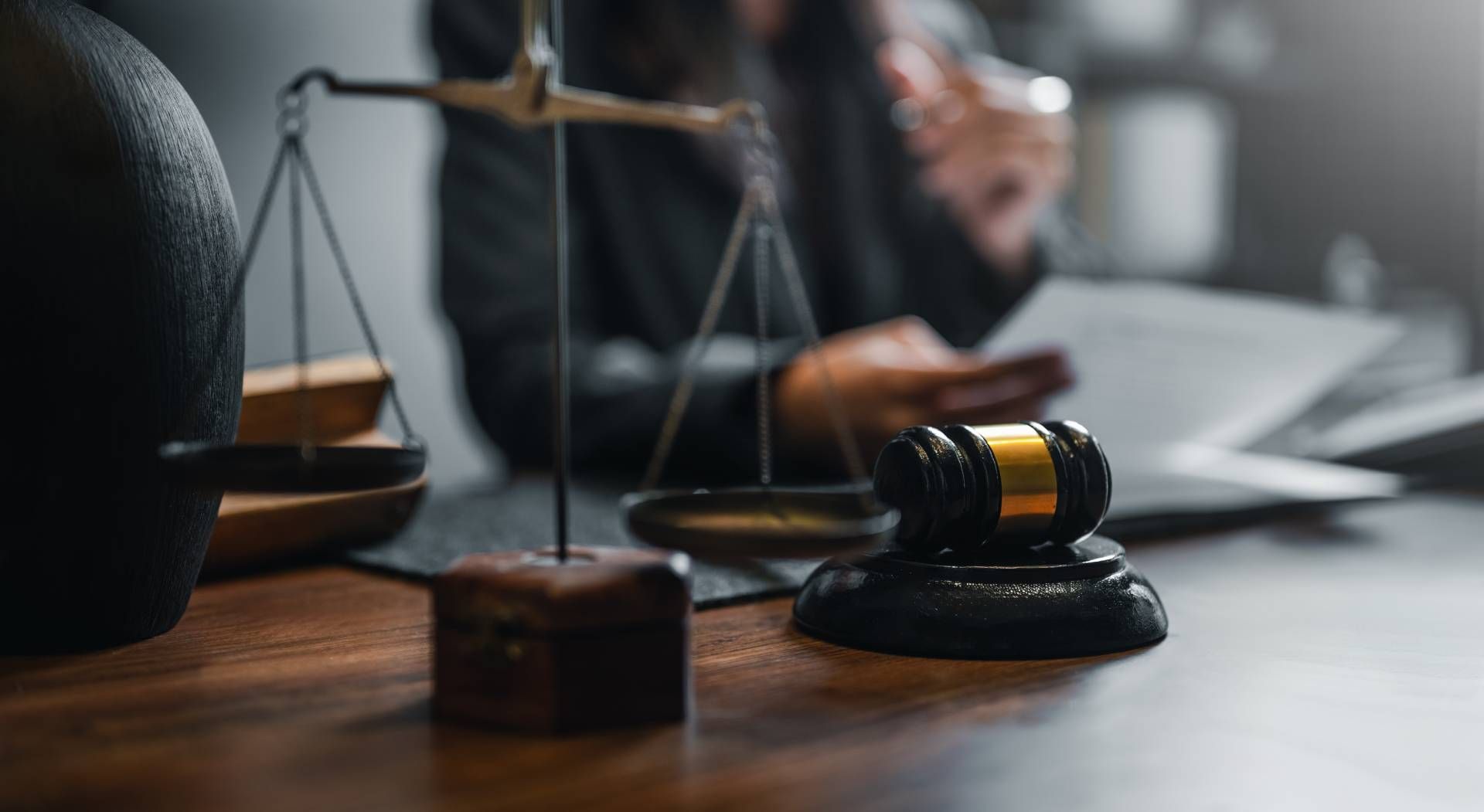 Gavel, scales of justice, and person in background holding documents at a desk, likely a legal setting.