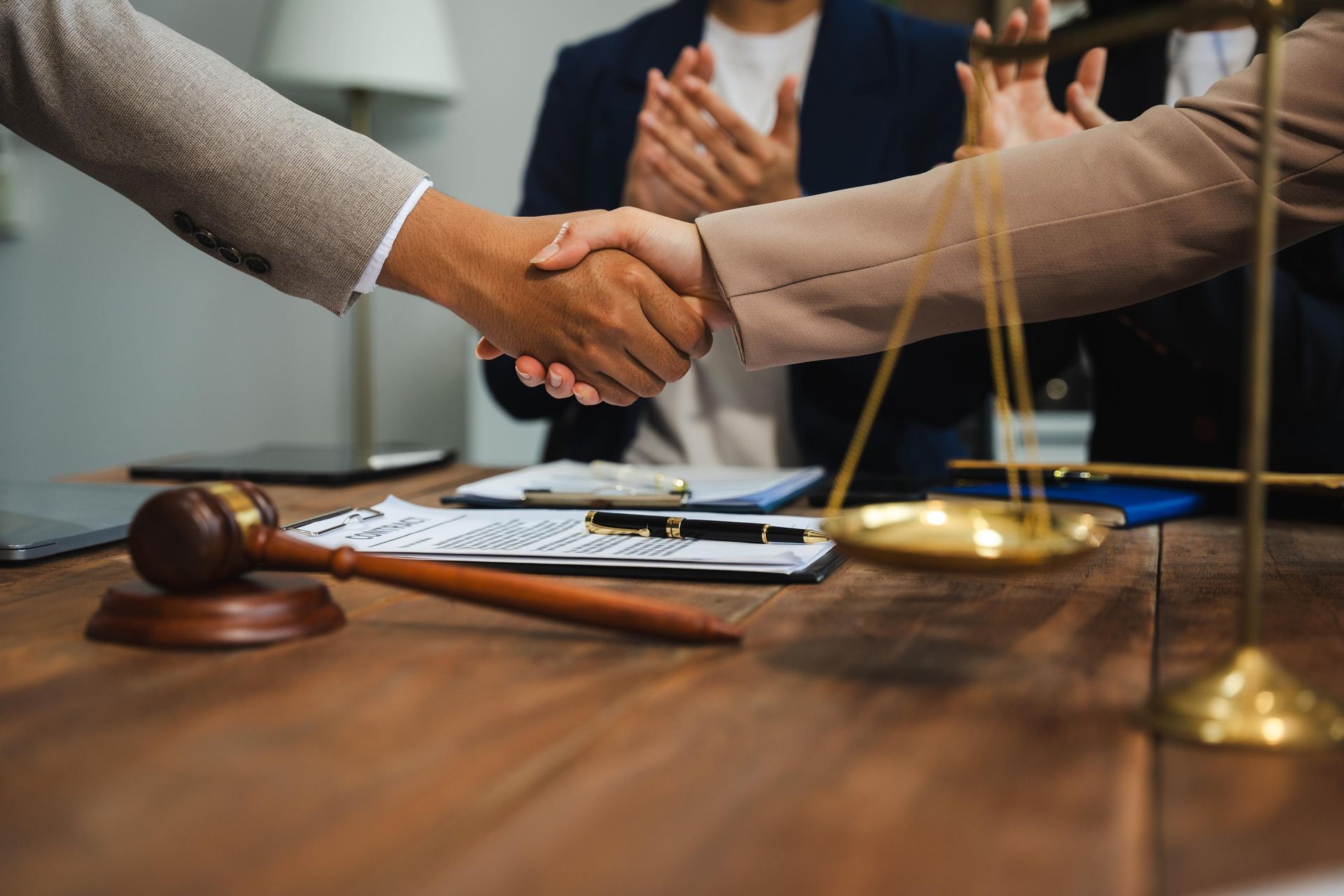 Two people shaking hands over a wooden table with a judge’s gavel and a scale of justice in the foreground.