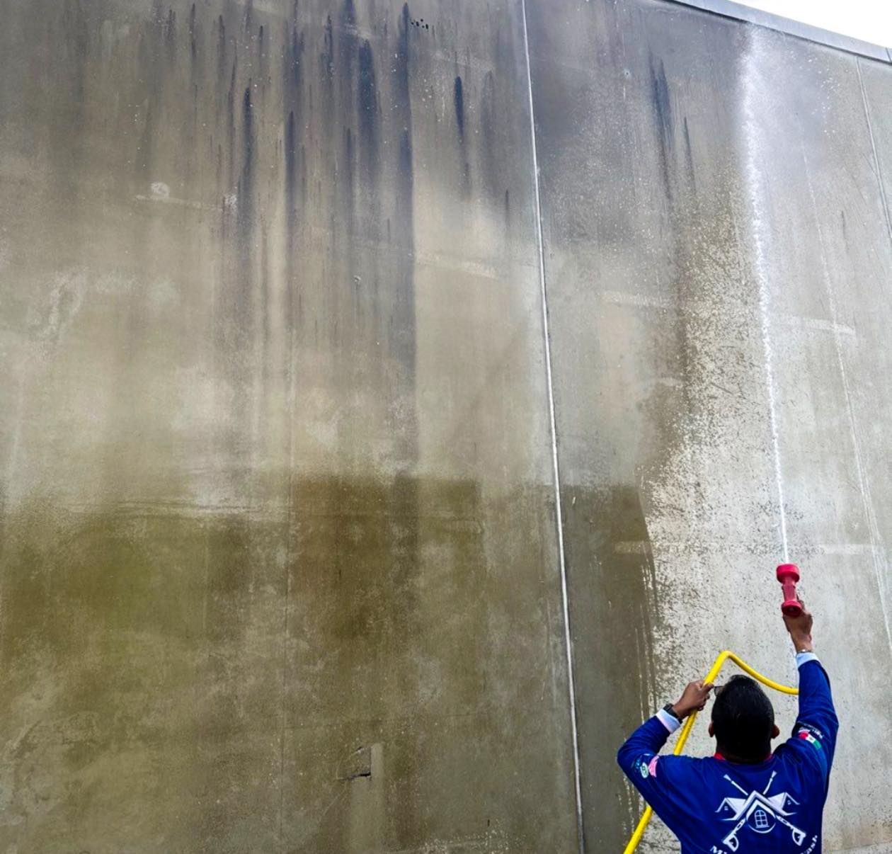 Worker pressure-washes a large concrete wall, spraying water upward with a red nozzle.