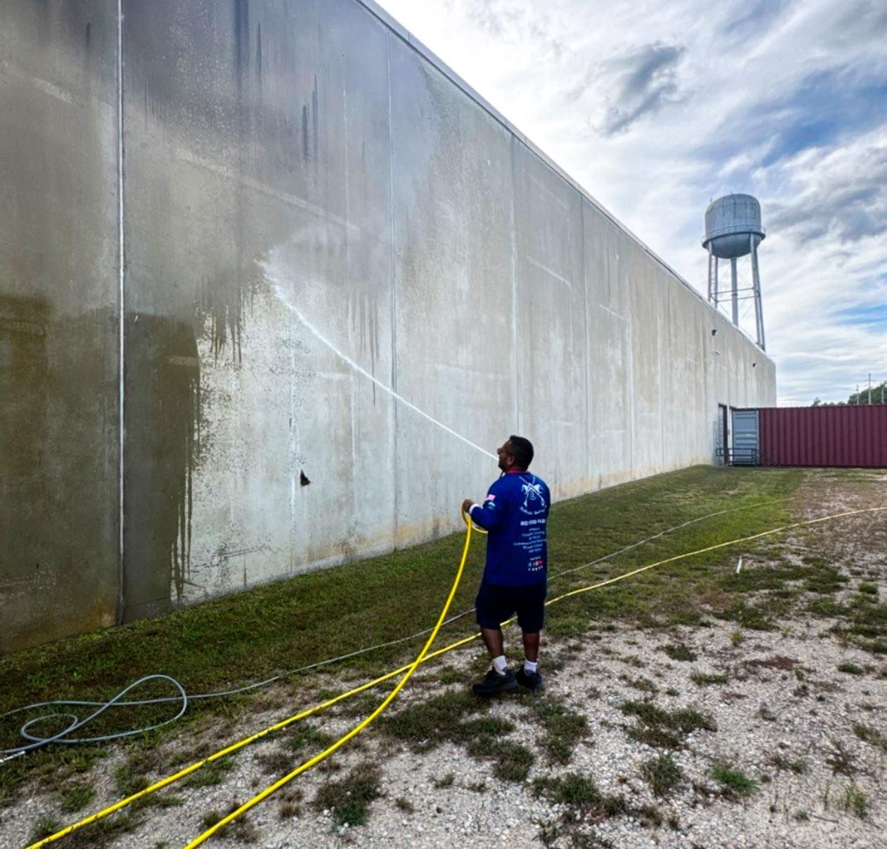 Person pressure-washing a large concrete wall beside a grassy yard and water tower.