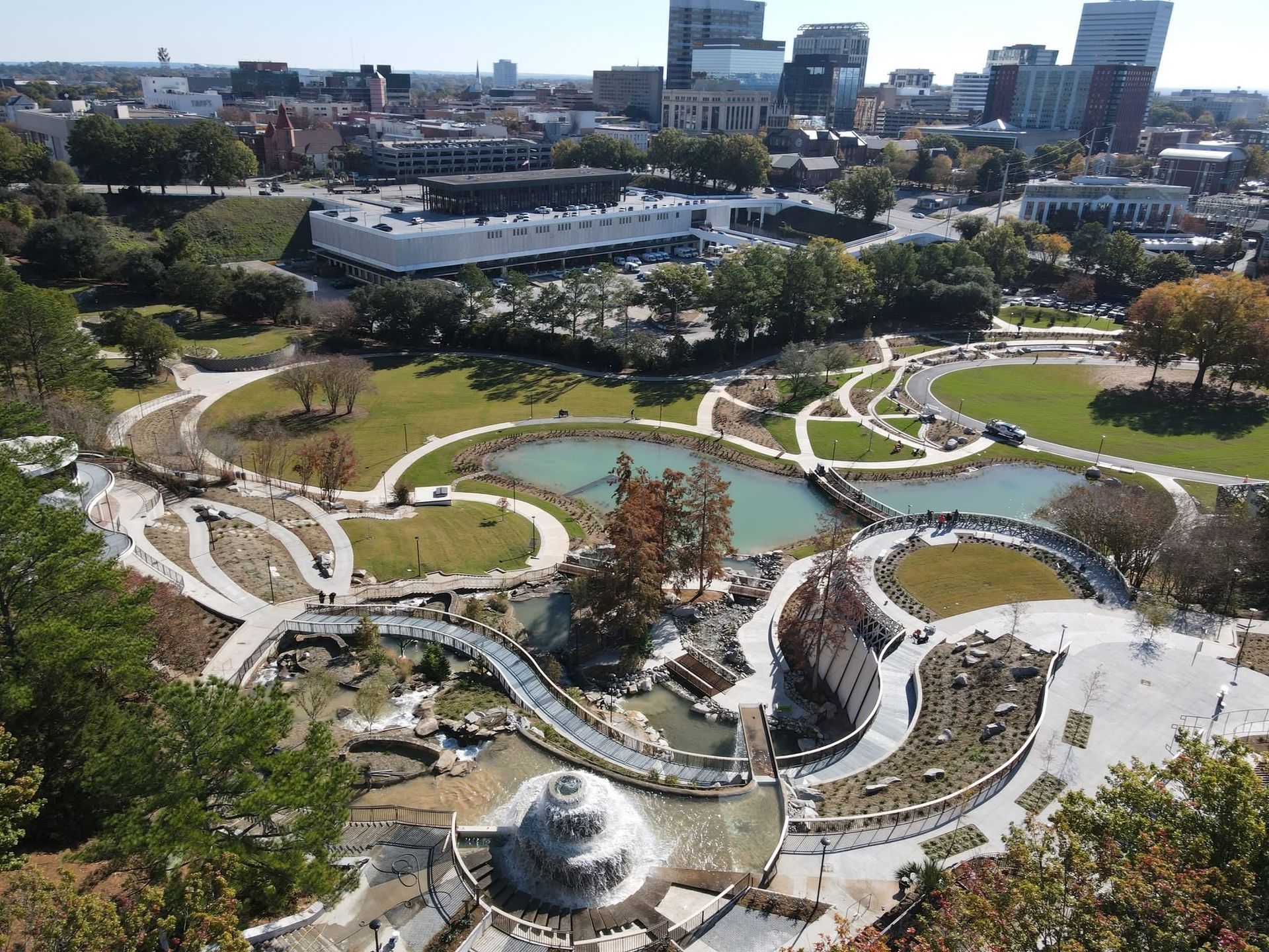Aerial view of a landscaped park with winding paths, ponds, and trees beside city buildings.