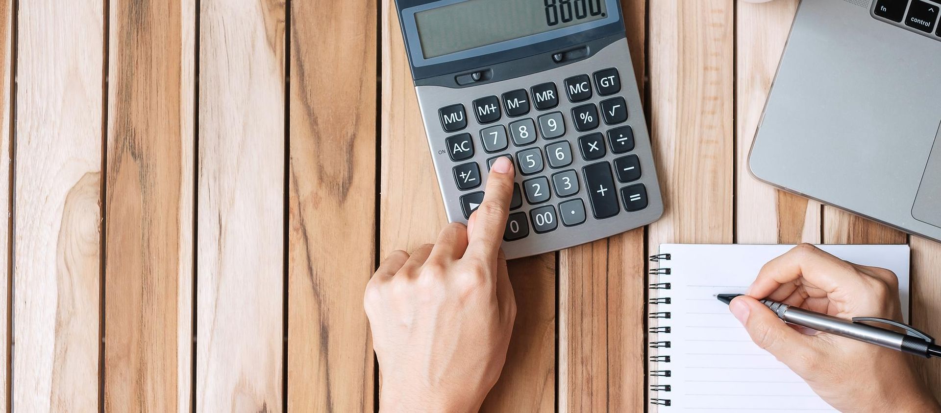 Hands using a calculator and notebook on a wooden desk with a laptop nearby