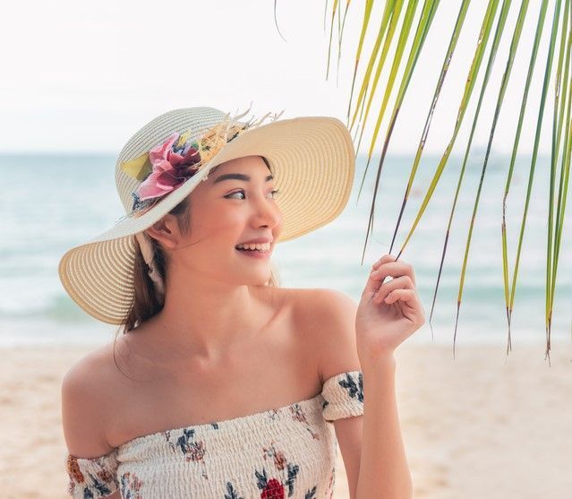 A woman wearing a hat is holding a palm tree branch on the beach.