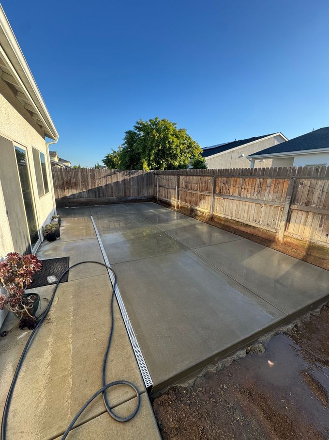 Backyard patio with wet concrete, hose, and wooden fence under bright blue sky