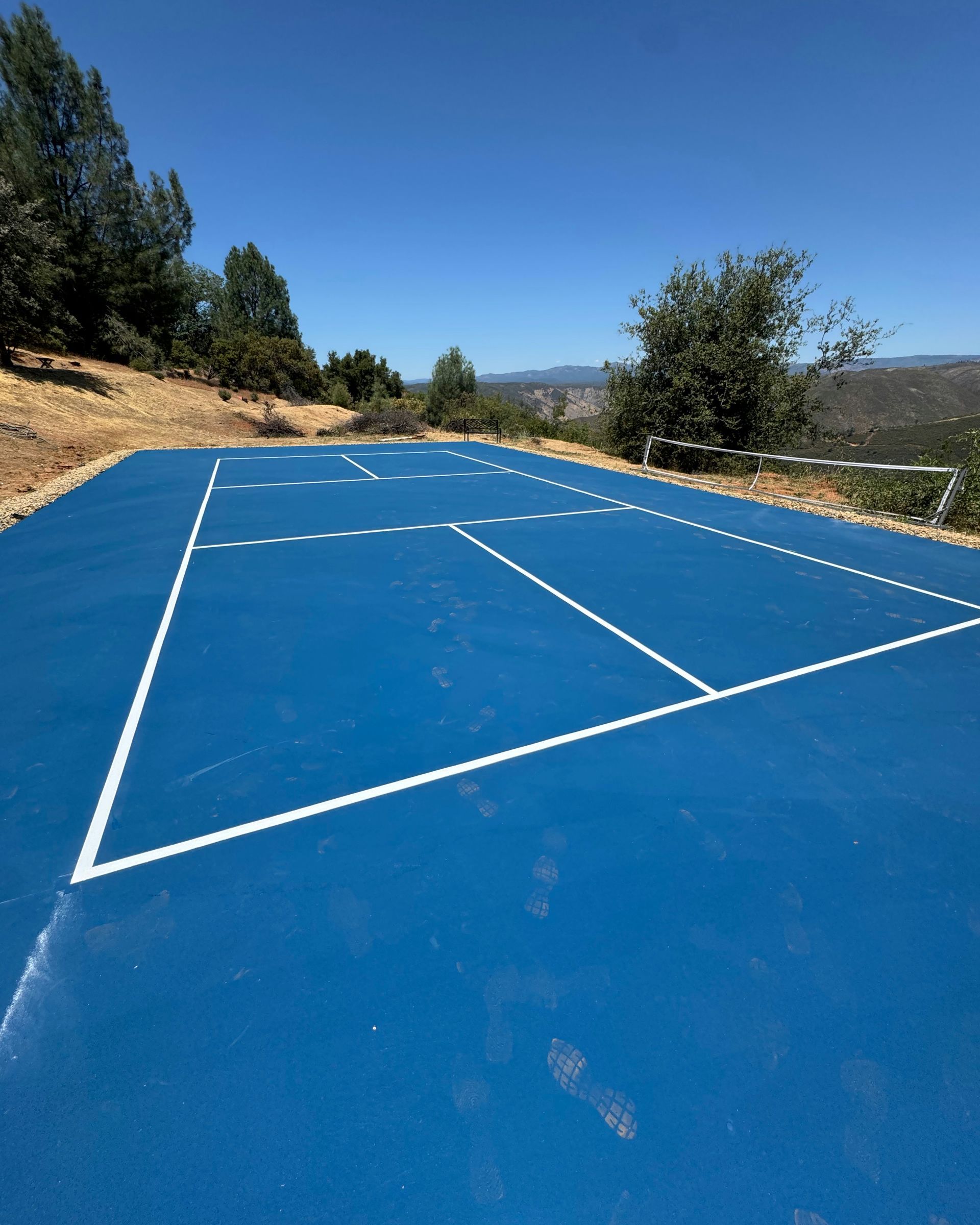 Blue outdoor court with white lines on a sunny hillside, bordered by trees and a distant valley.