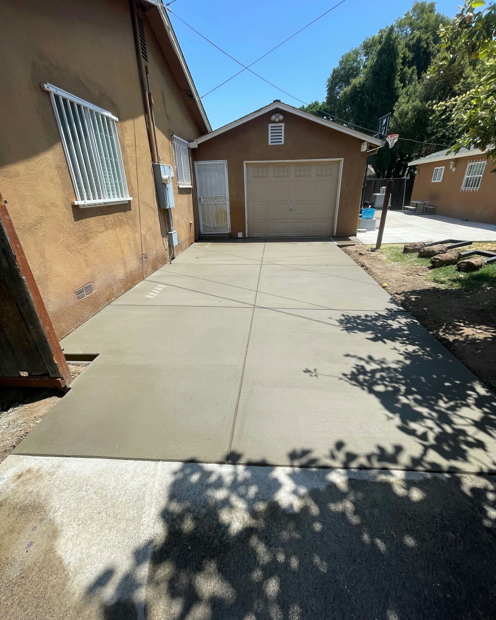 Concrete driveway leading to a tan garage on a sunny residential property, with trees casting shadows.