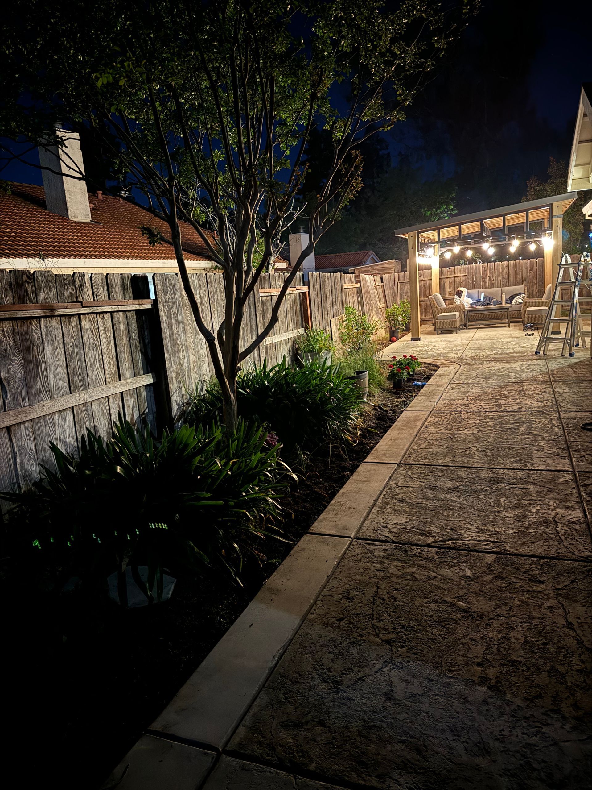 Nighttime garden walkway beside a lit patio with a tree, fence, and house lights.