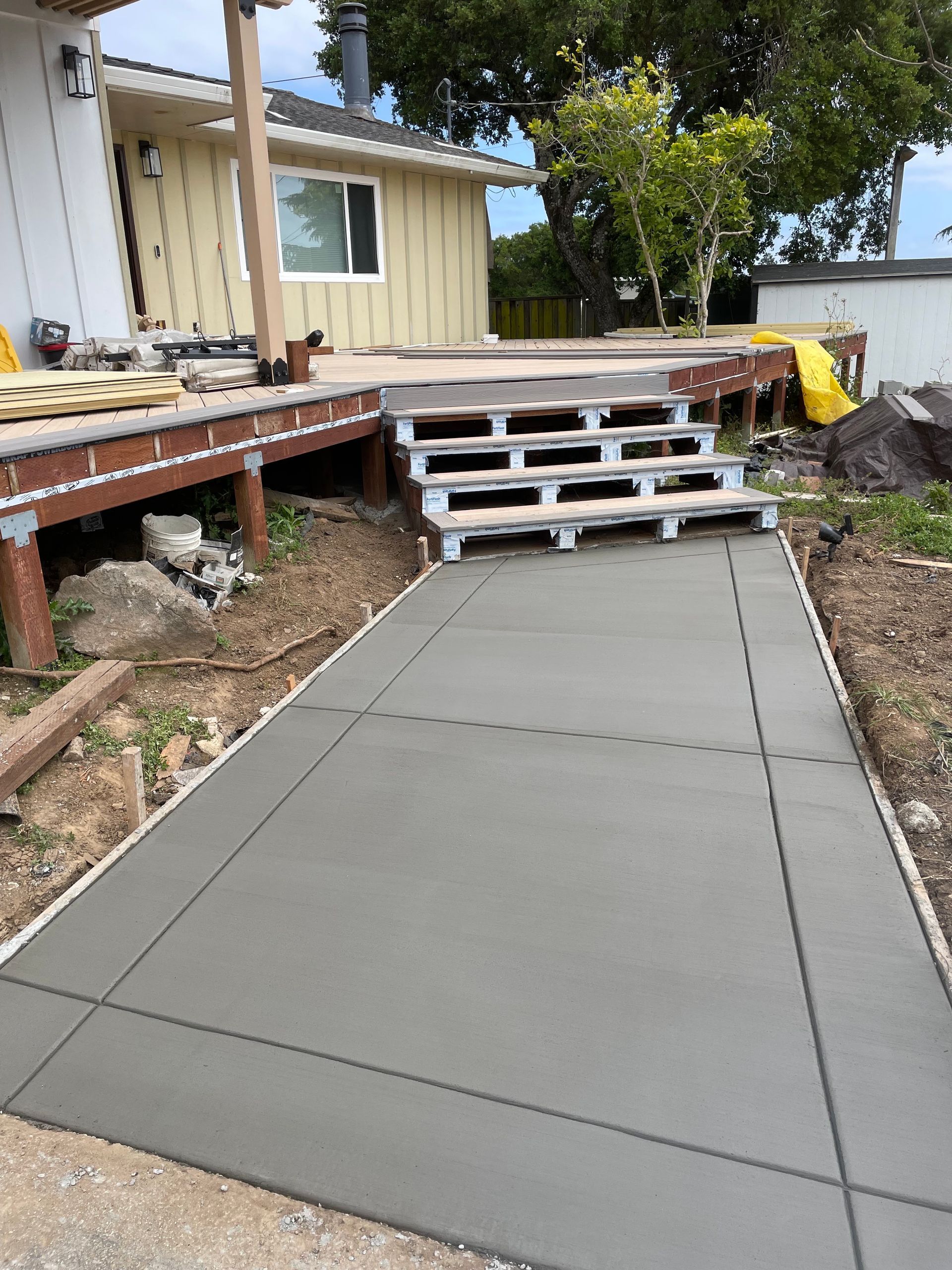 Fresh concrete walkway leading to a raised wooden deck beside a beige house