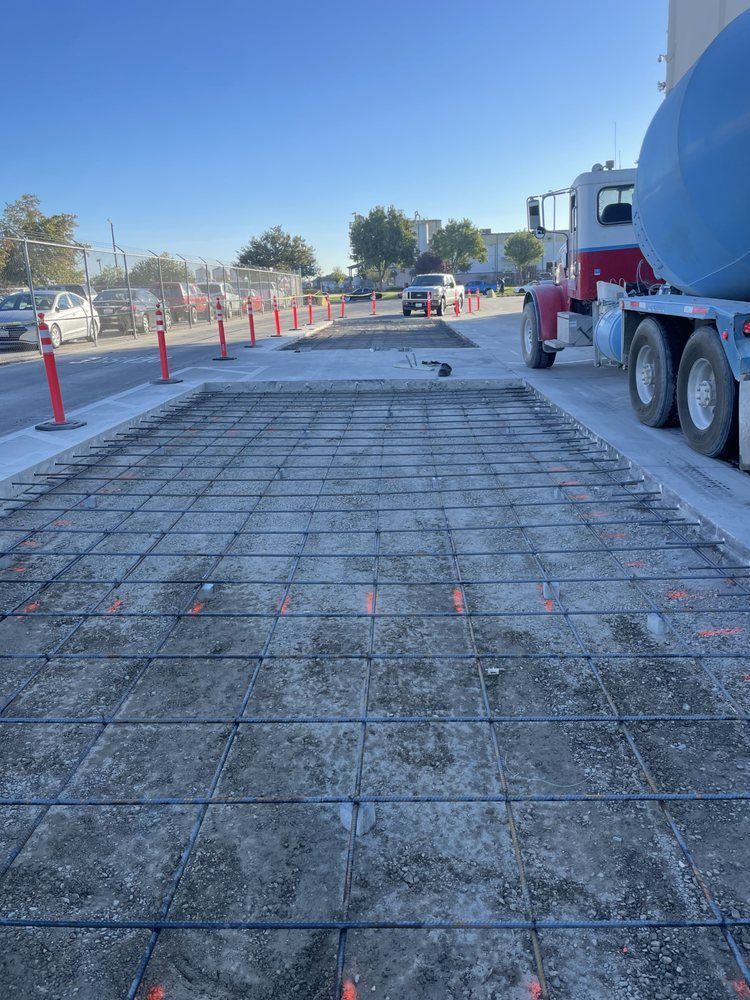 Construction site with rebar grid slab, orange cones, and a concrete mixer truck on a road under clear blue sky