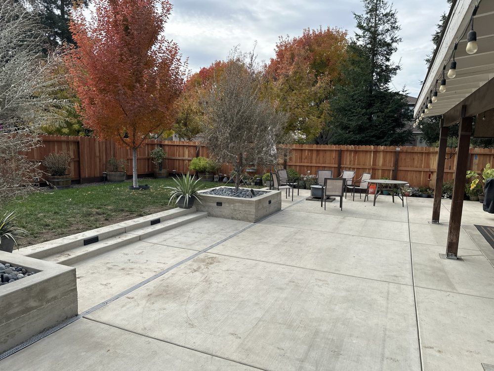Backyard patio with concrete slabs, raised planters, chairs, and autumn trees behind a wooden fence